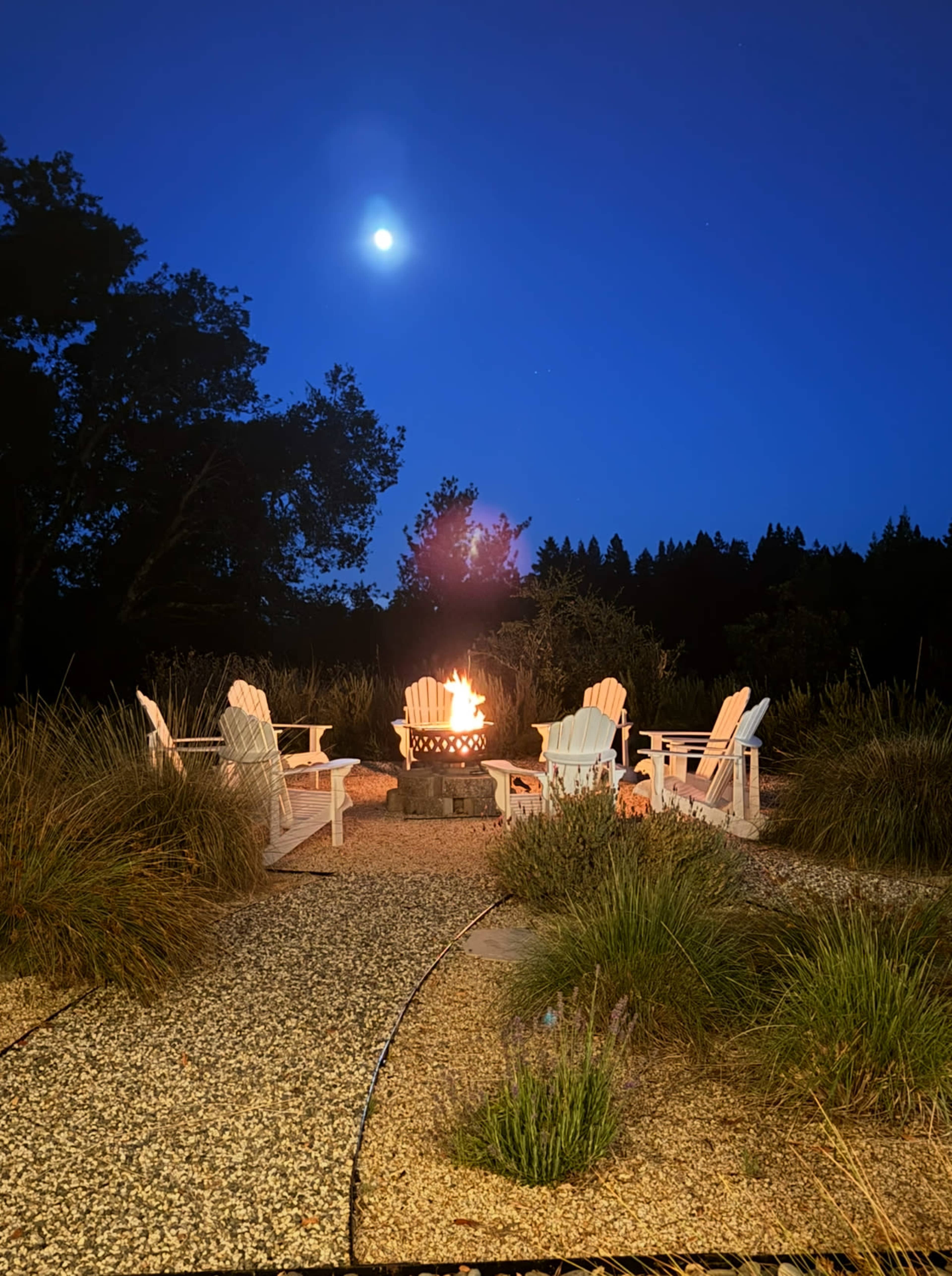 A stone fire pit surrounded by white Adirondack chairs sits on a gravel path under a clear night sky with a bright moon.