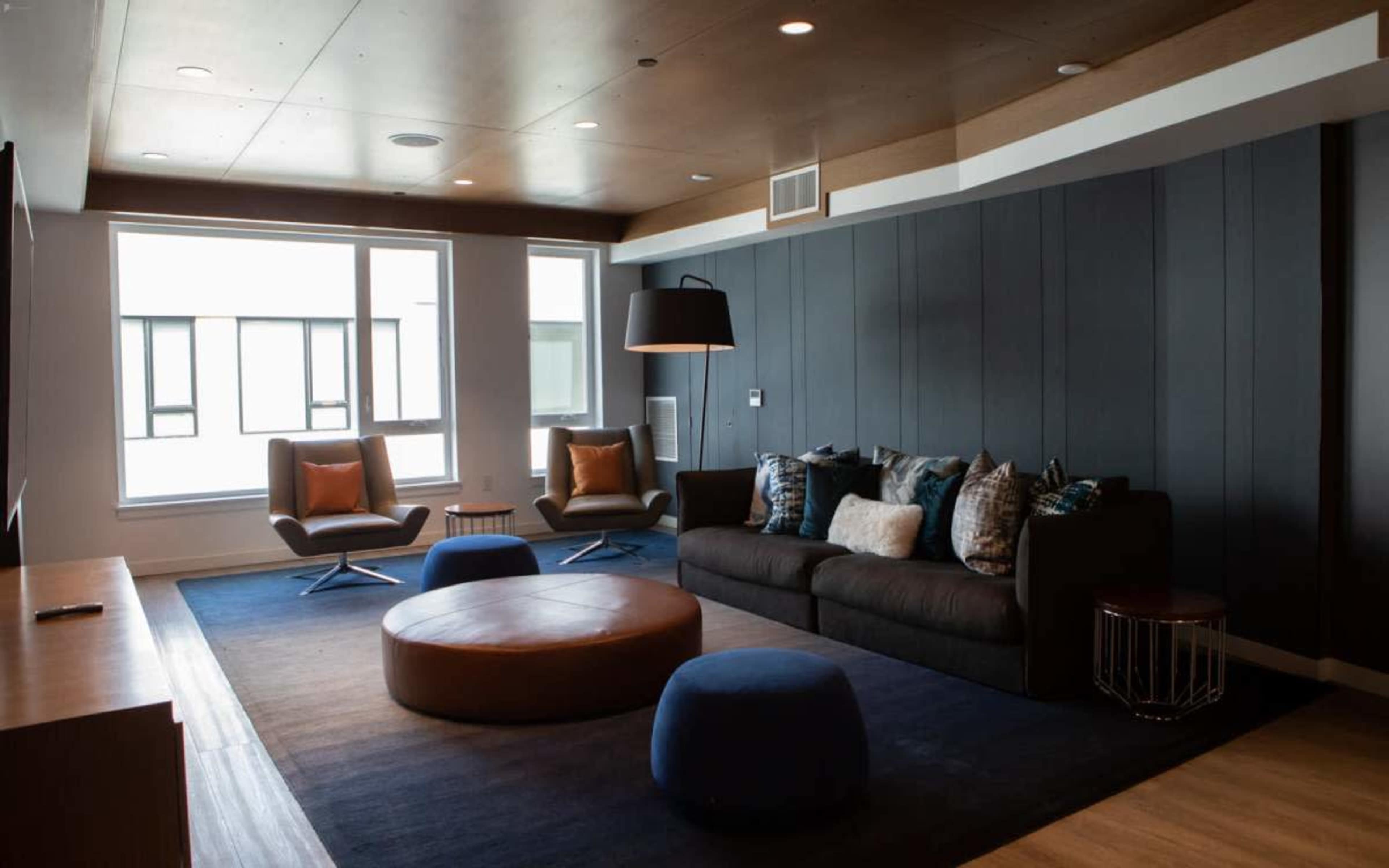 The image shows a modern living room featuring a dark sofa, two lounge chairs, a round ottoman, and large windows.