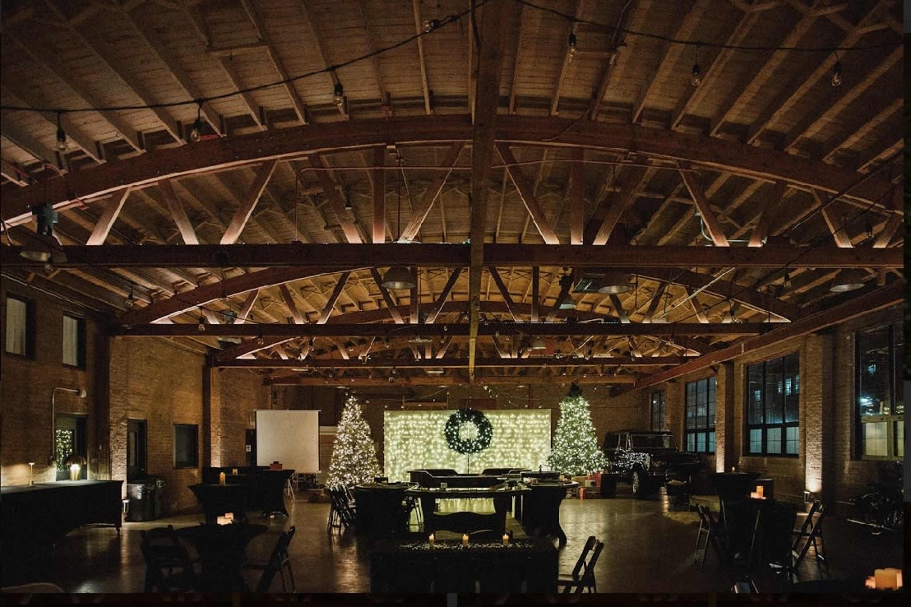 A spacious event venue features wooden beams, festive decorations, and tables arranged for a gathering, with Christmas trees and lights adorning the space.