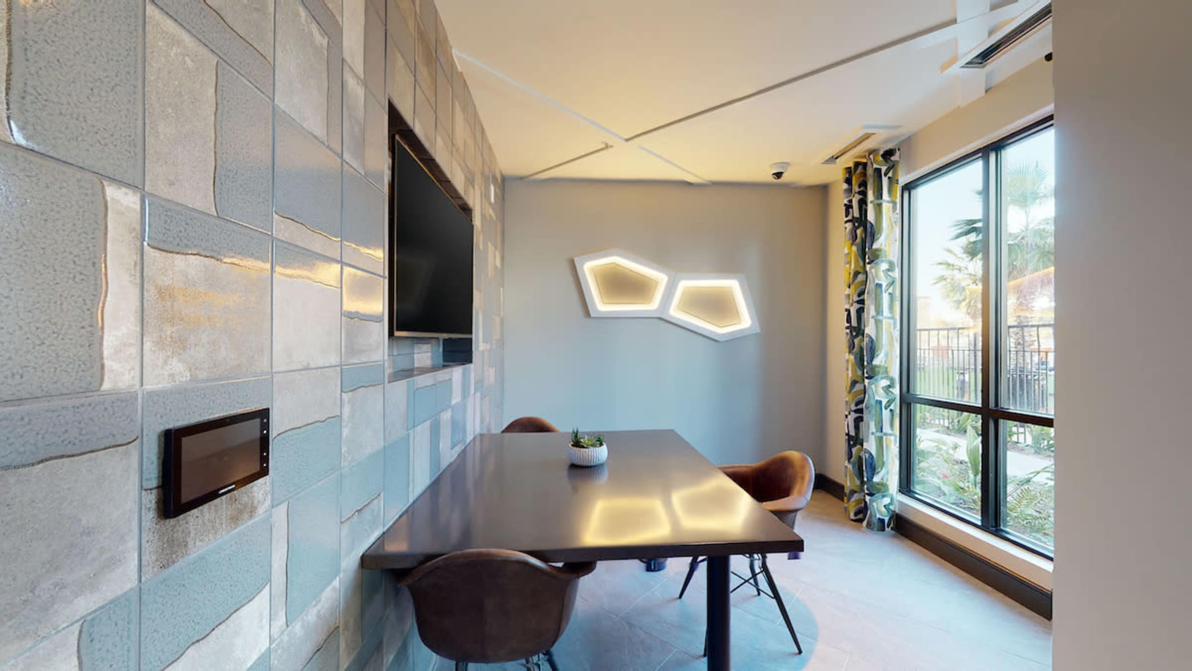 The image shows a modern conference room with a rectangular table, four brown chairs, a wall-mounted television, and decorative wall tiles.