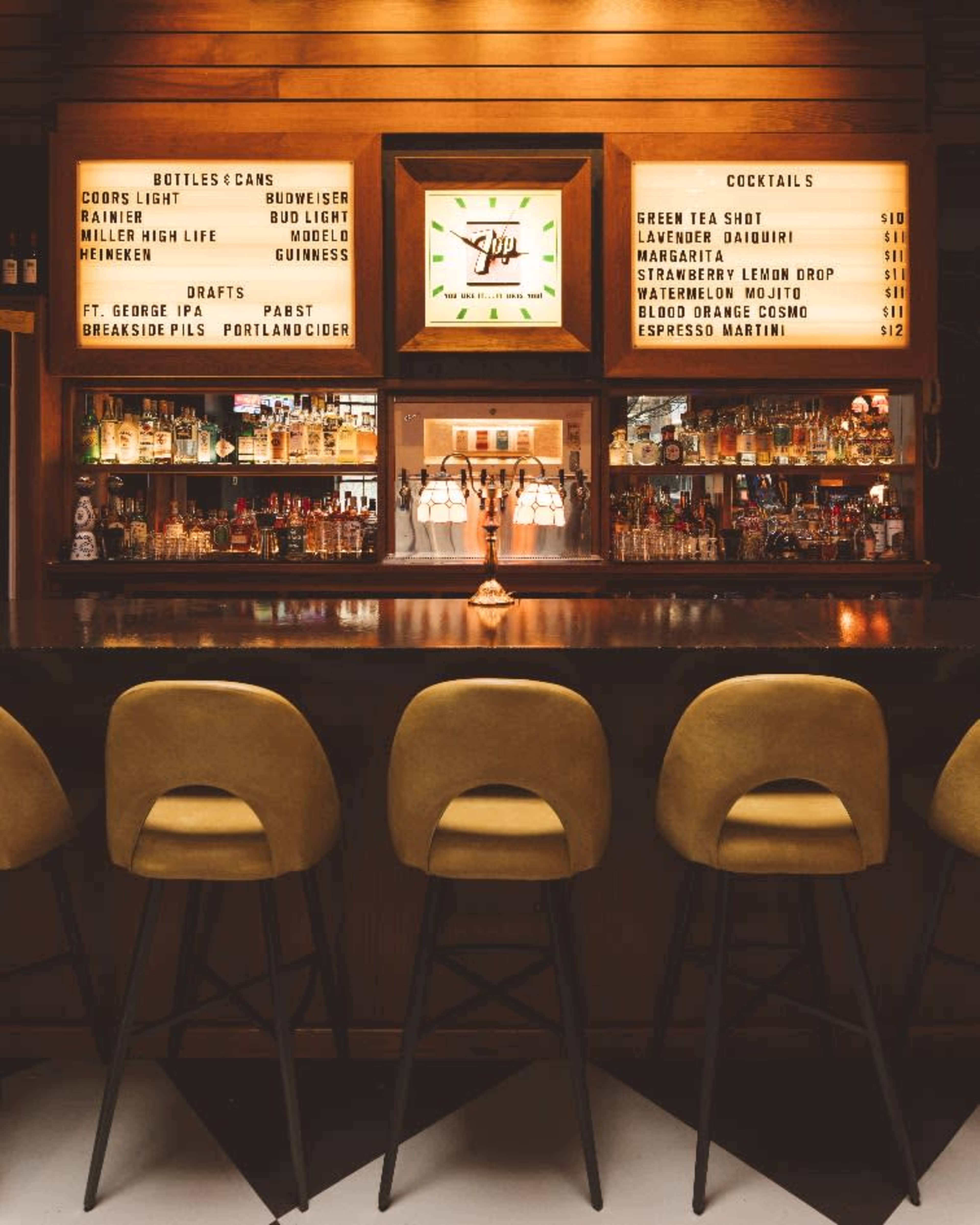 The image shows a well-lit bar with a wooden back wall, displaying various bottles and a menu of cocktails.