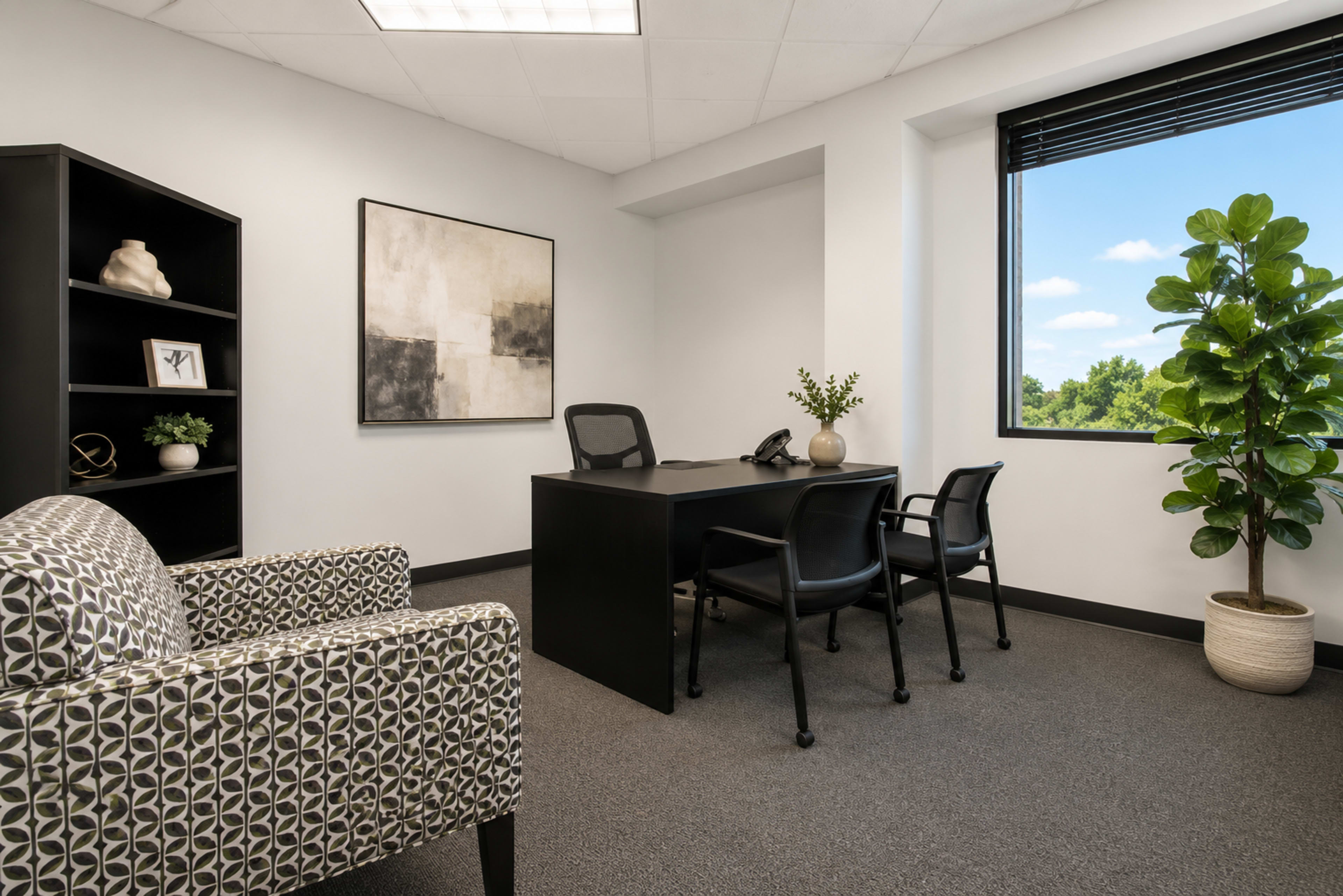 The image shows a modern office space furnished with a black desk and three chairs, a patterned armchair, a black bookshelf, and a large window overlooking greenery.