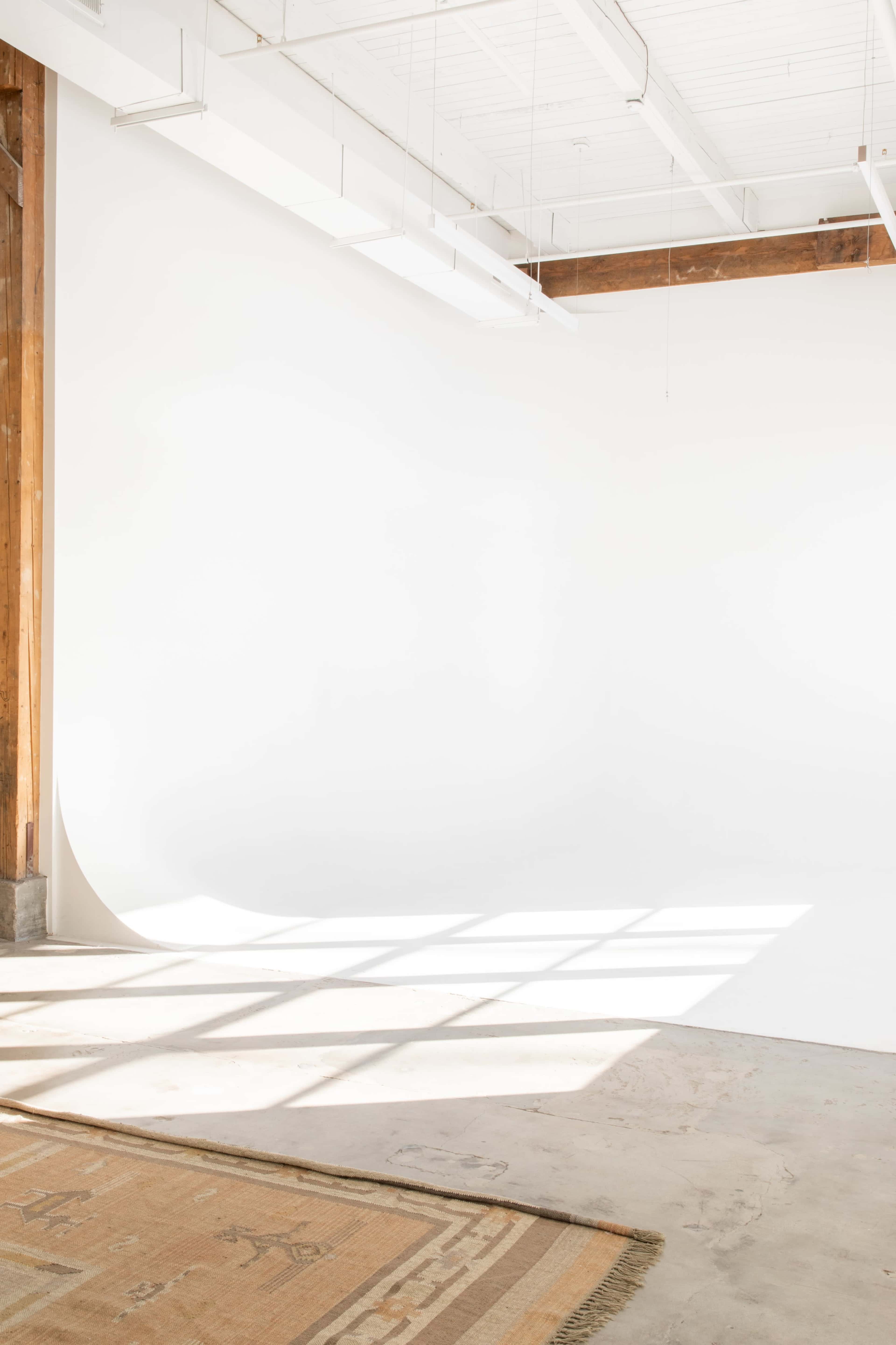 The image shows a spacious interior with a large white backdrop, wooden beams, and a patterned area rug on the floor.