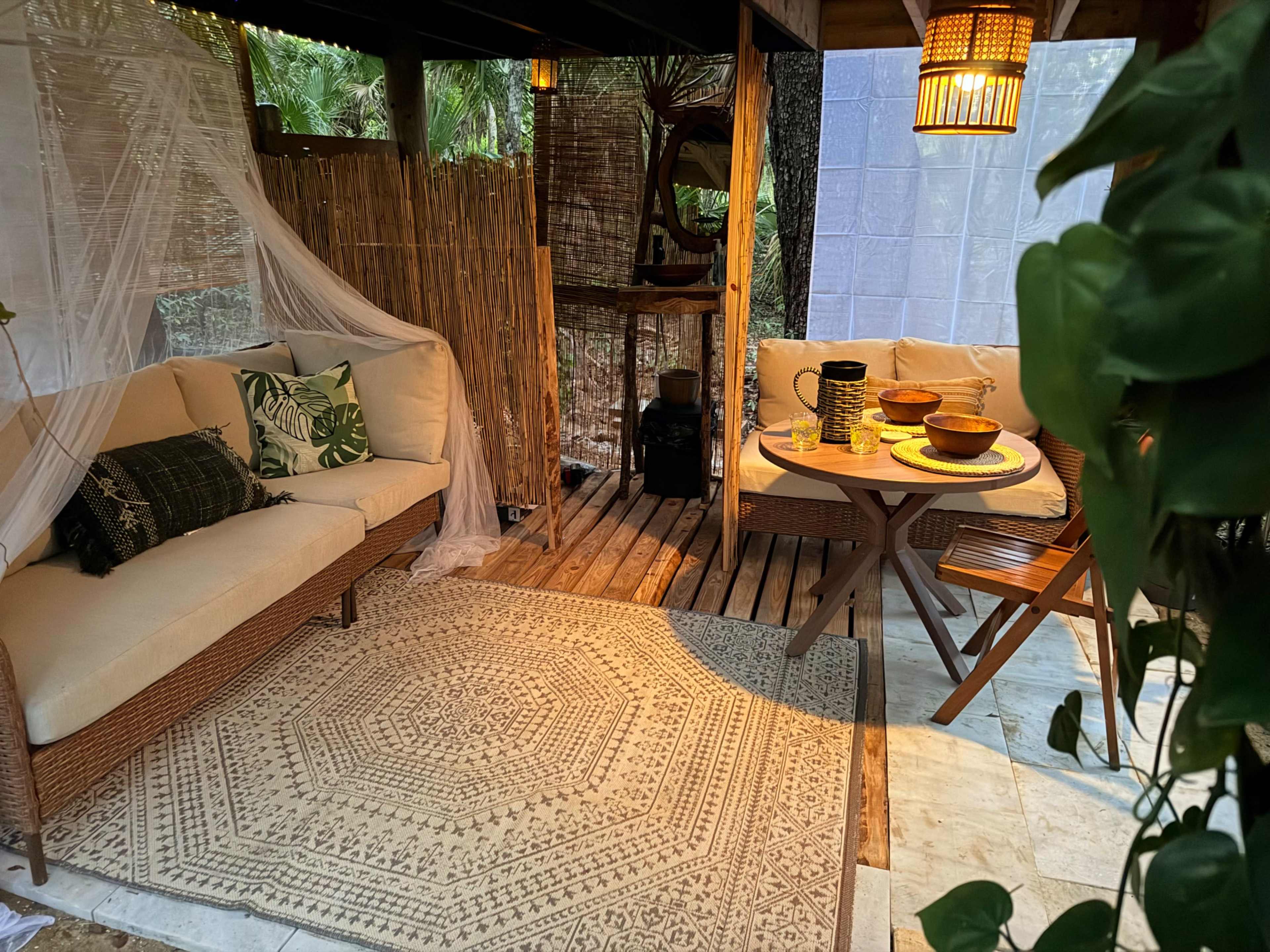 The image shows a cozy outdoor seating area with a woven sofa, a wooden dining table, and decorative lanterns, surrounded by bamboo and plants.