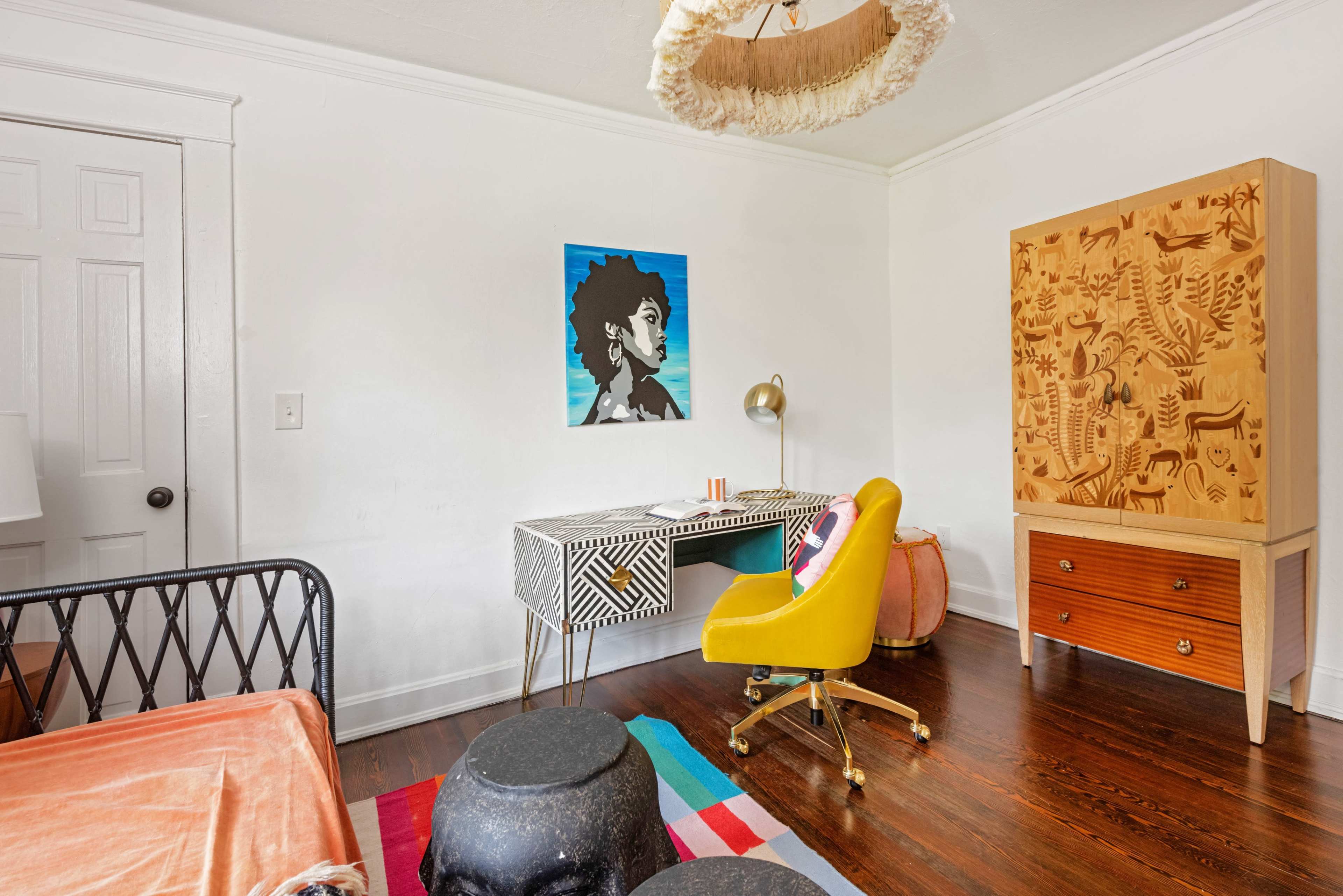 The room features a bed, a decorated wall with a colorful portrait, a desk with a yellow chair, and an ornate wooden cabinet.
