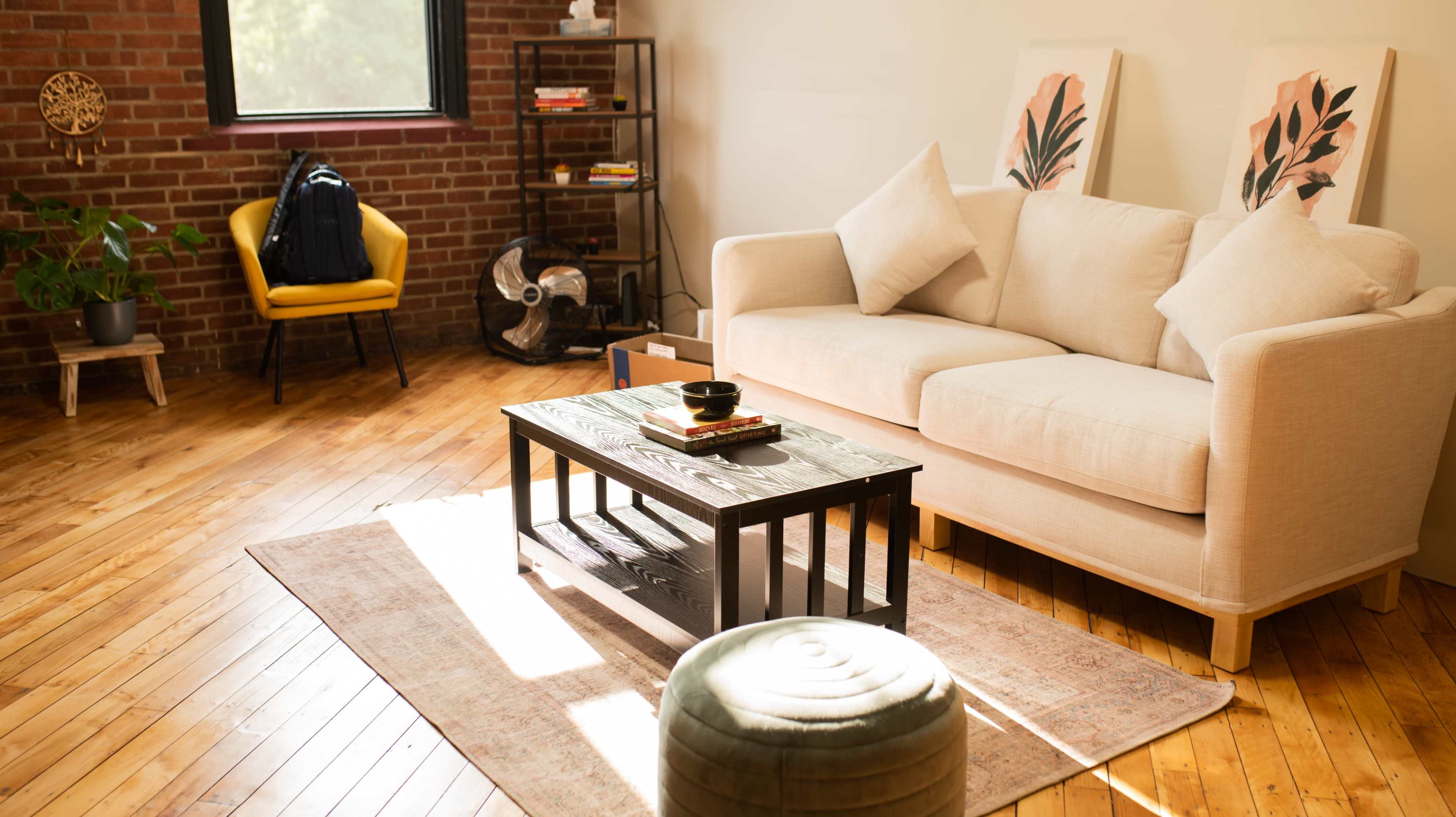 A cozy living space featuring a light-colored sofa, a black coffee table, a patterned rug, and a yellow chair, with a backpack and a decorative plant visible in the background.
