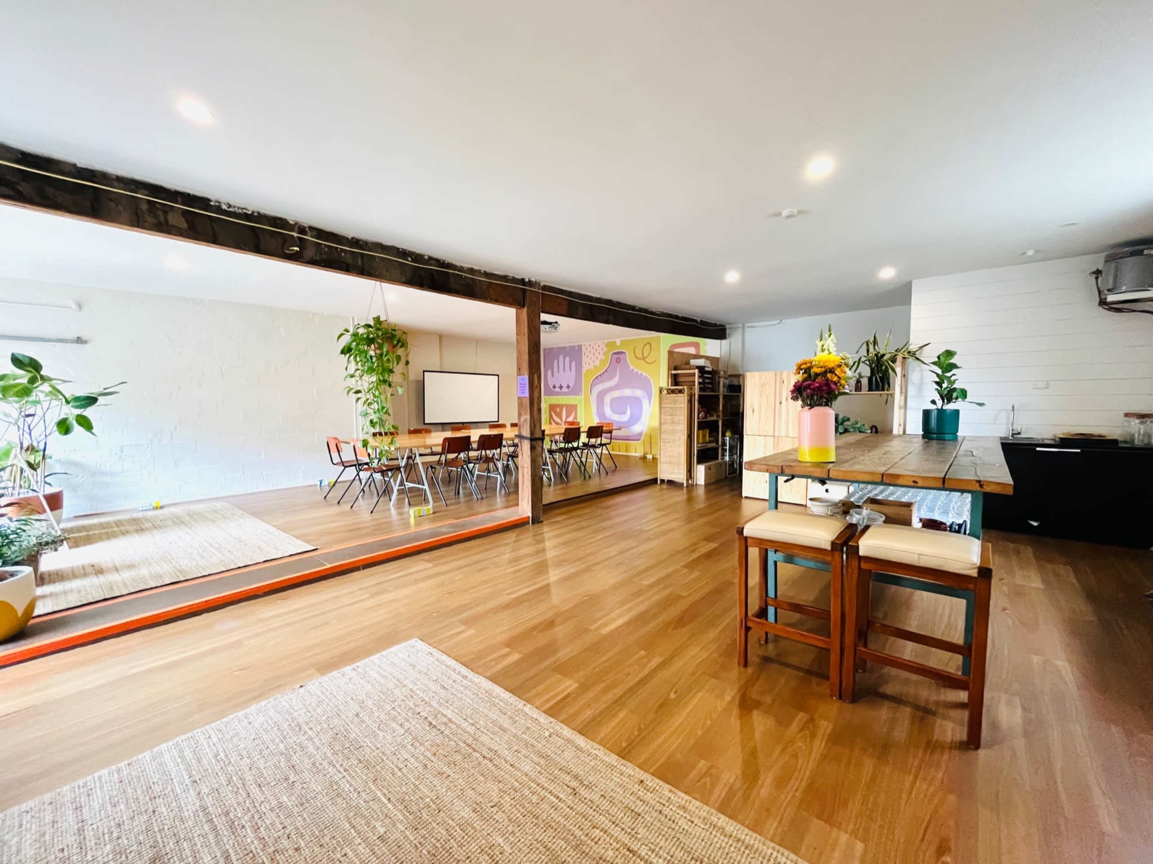The image shows a spacious room with wooden flooring, featuring a dining area with a long table and chairs, plants, and a partition with a colorful design.