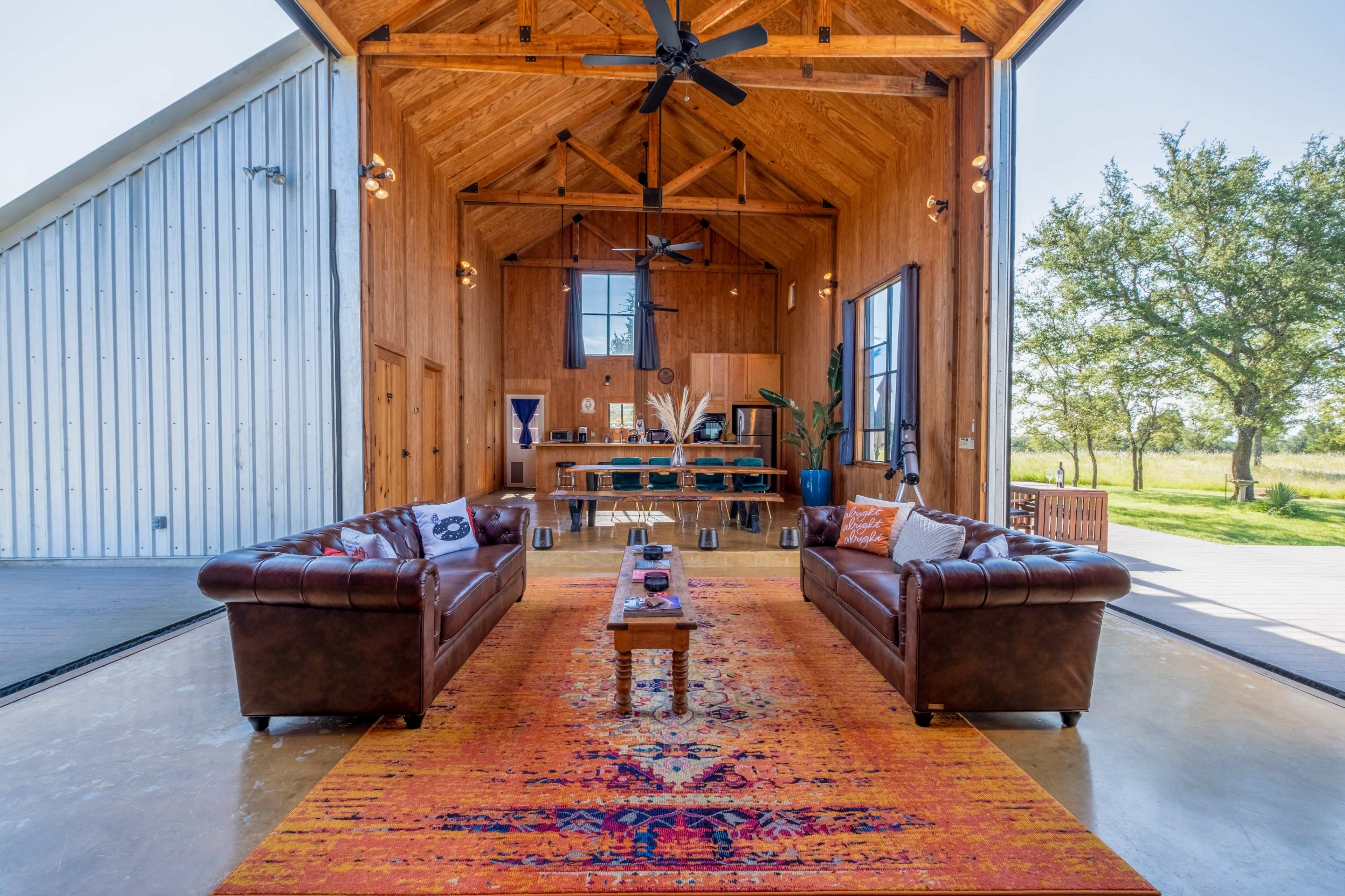 The image features a spacious, modern interior with wooden walls and a high ceiling, furnished with two brown leather sofas facing a central coffee table on a vibrant area rug, and a dining area visible in the background through large open doors.