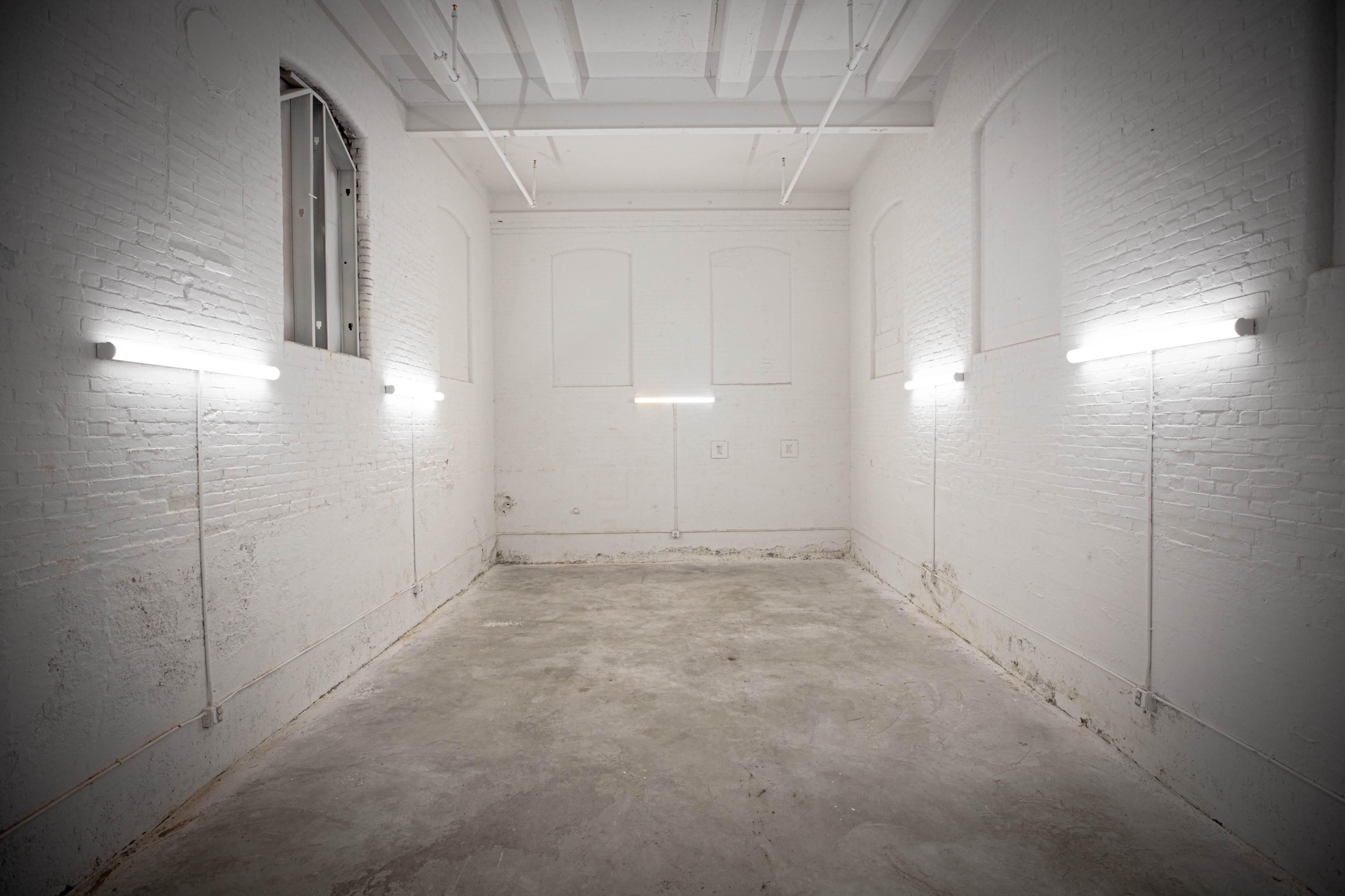 An empty, unadorned room with white brick walls and overhead fluorescent lights illuminating the concrete floor.