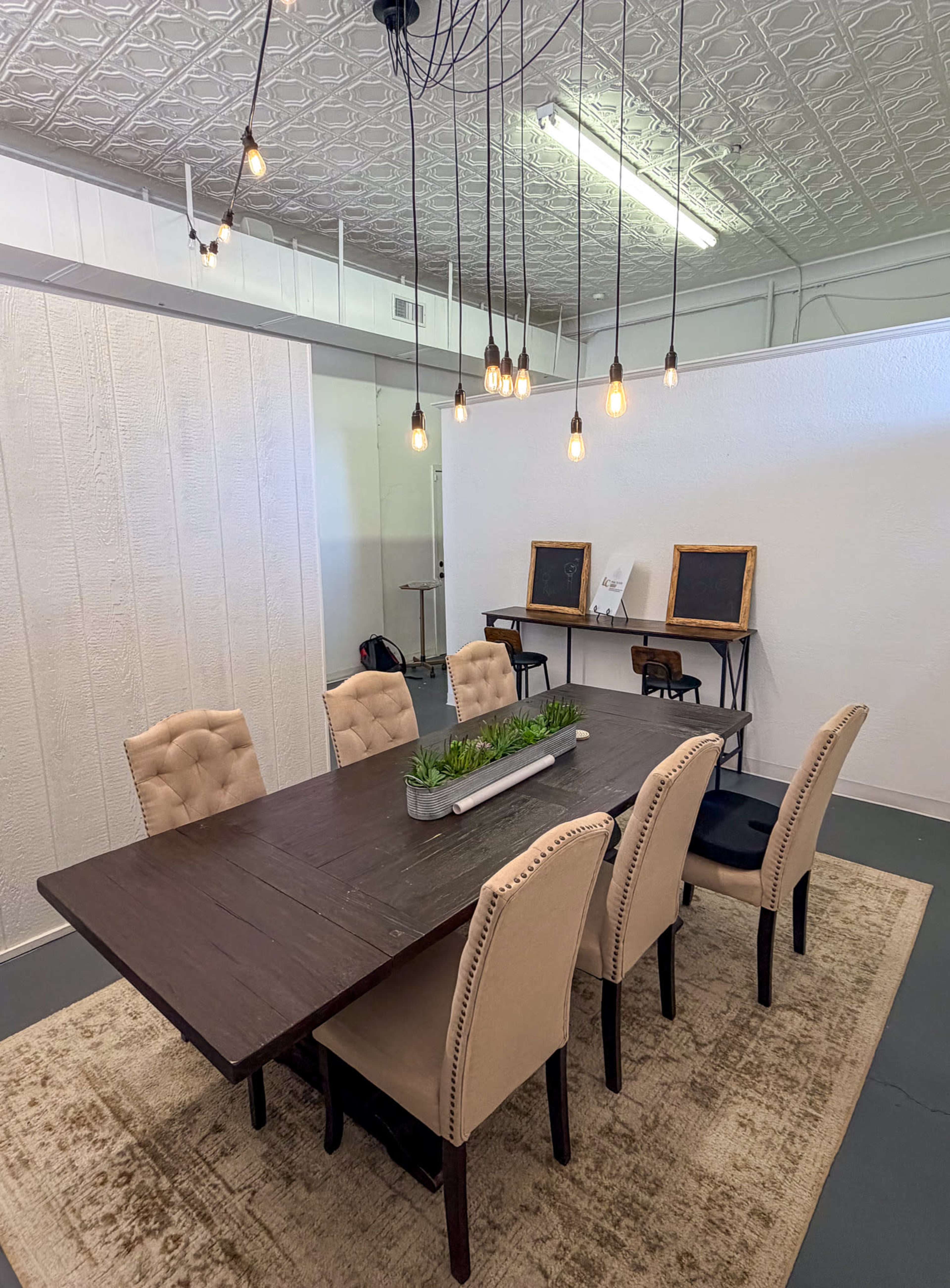 A long wooden table surrounded by upholstered chairs is set in a room with a textured ceiling and illuminated by hanging light bulbs.