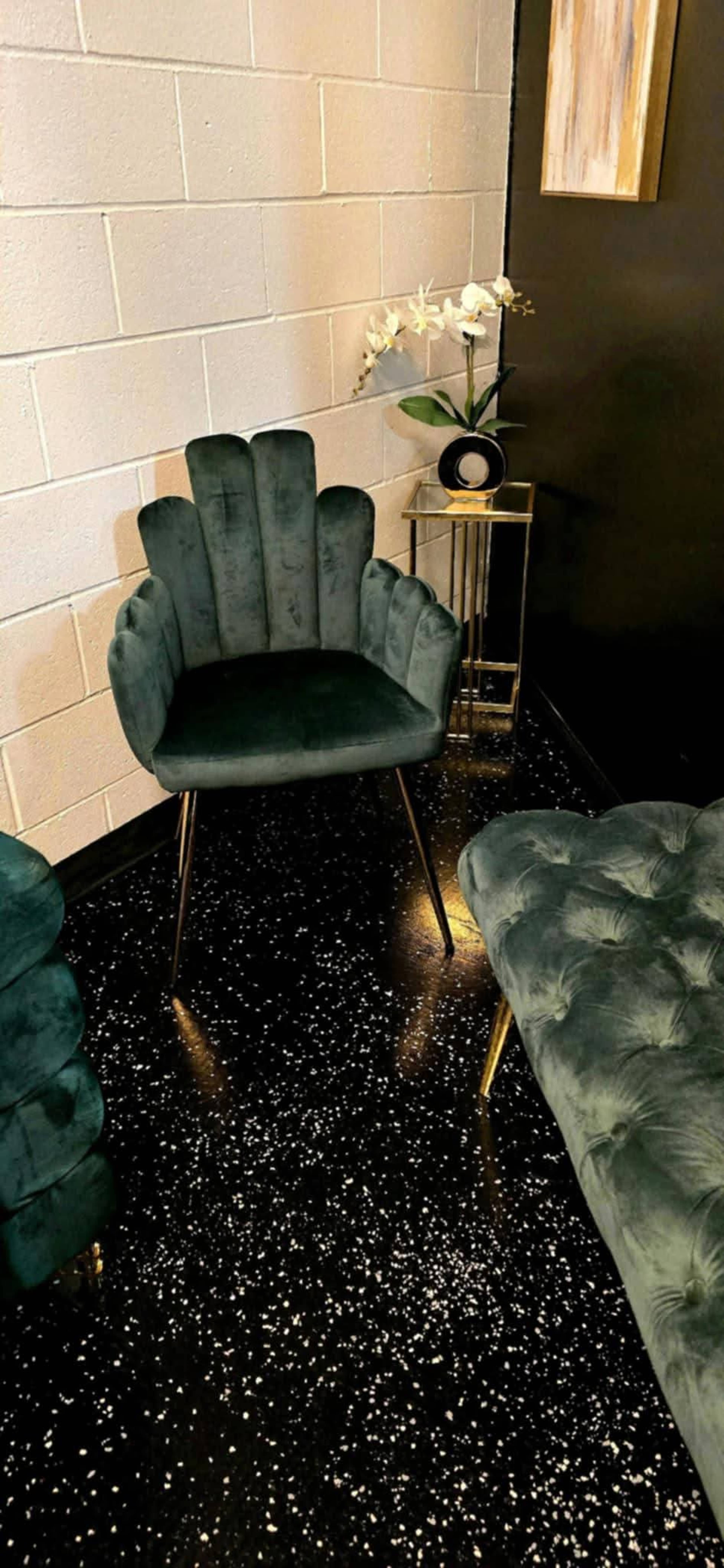 A plush, green velvet chair with a scalloped backrest is positioned next to a small table and an orchid plant in a dimly lit corner.
