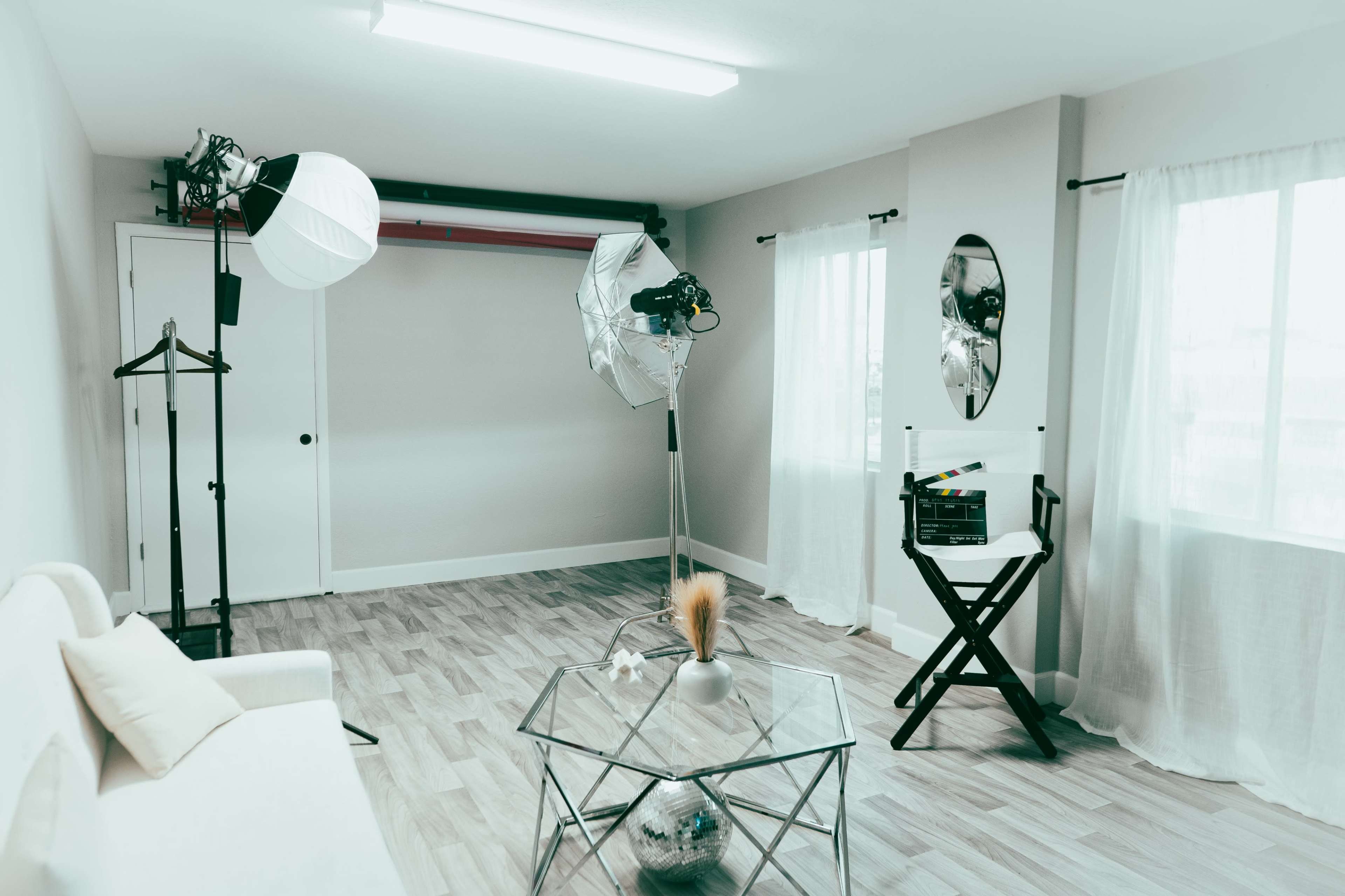 A bright photography studio features a white couch, a standing mirror, and two large lights set up beside a director's chair and a glass coffee table.