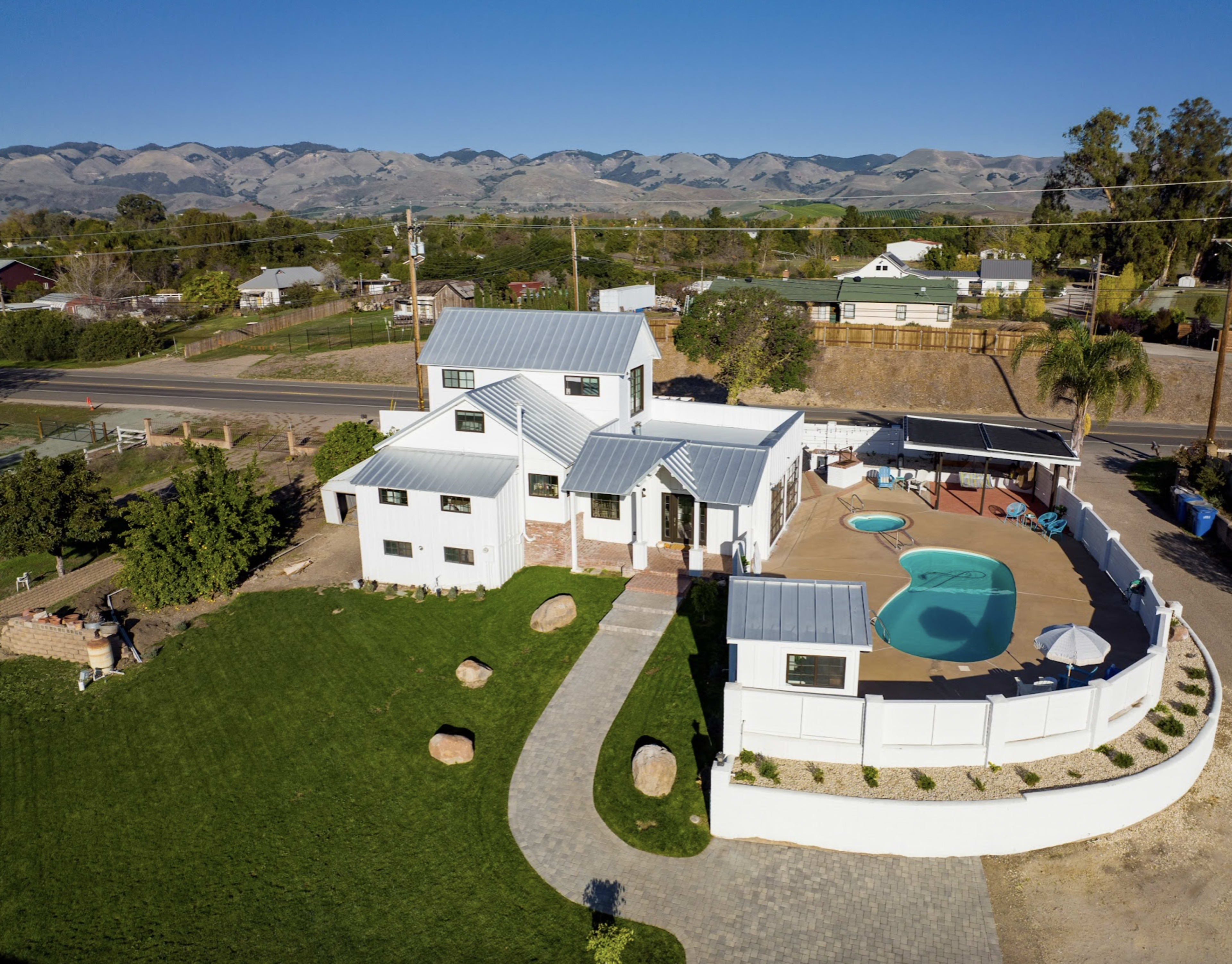 Aerial view of a white house with a grey metal roof, accompanied by a circular pool and a patio area, set against a backdrop of mountains.