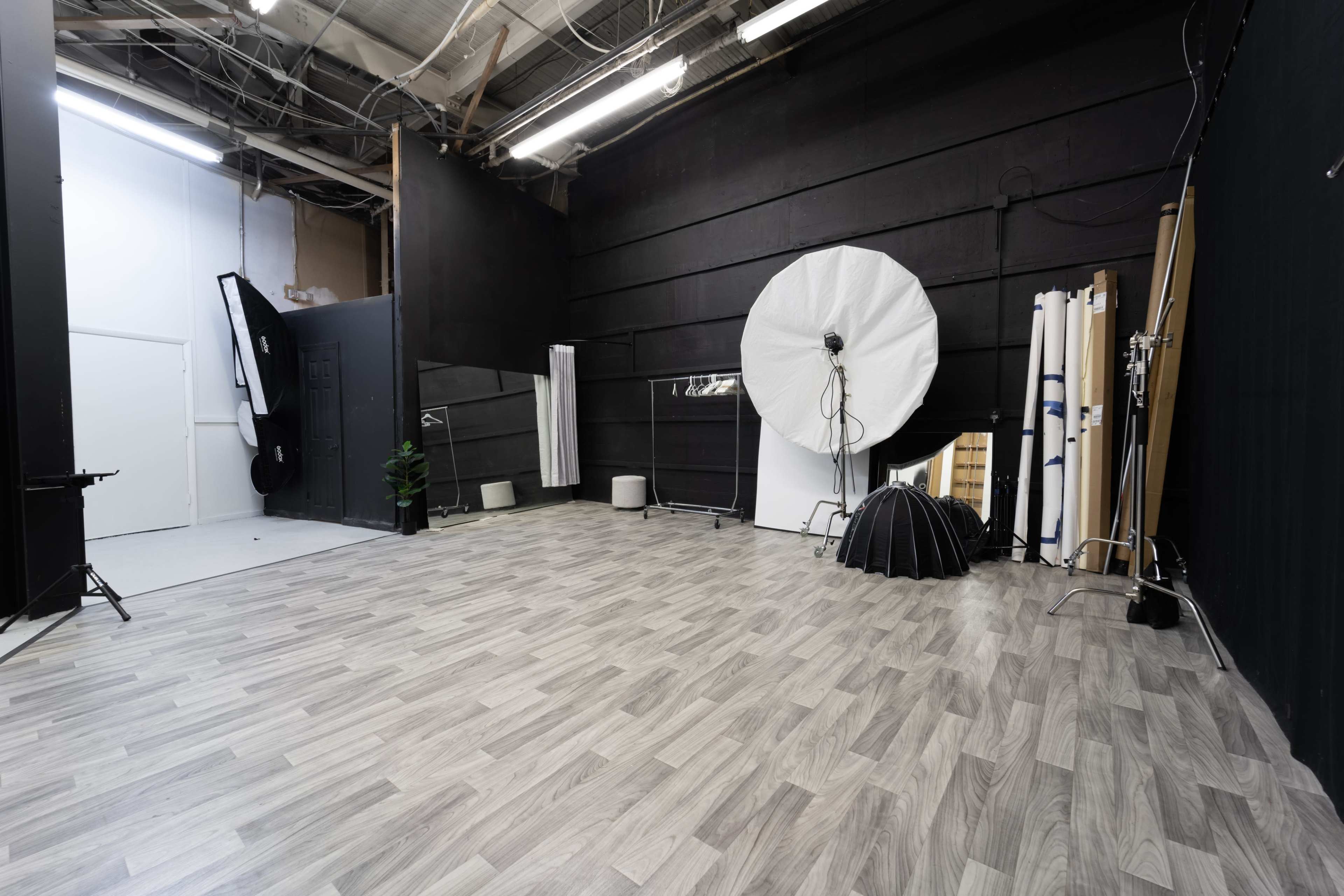 An empty studio space featuring a light-colored floor, black walls, and various photography equipment, including a large softbox and backdrop stands.