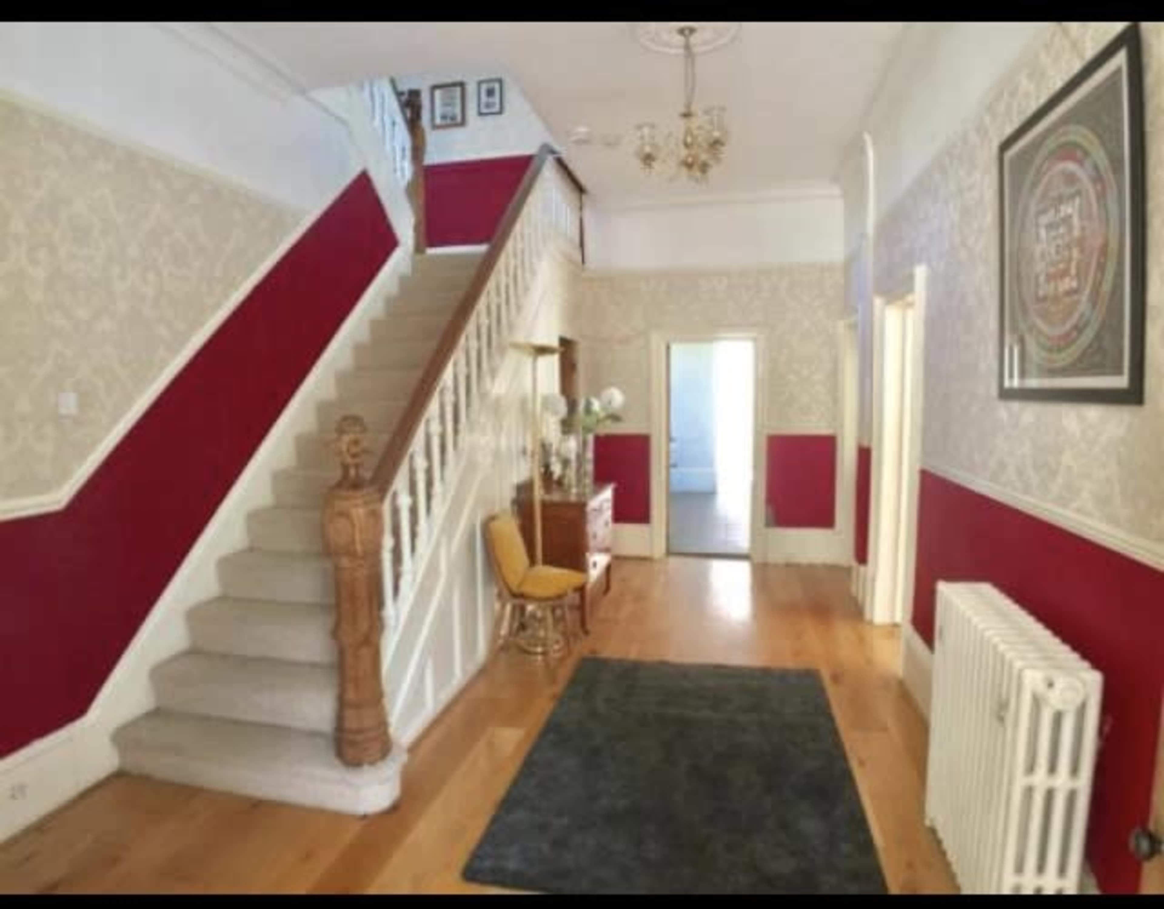 A spacious hallway with a staircase, featuring patterned wallpaper and a wooden floor, along with a decorative chair and a radiator.