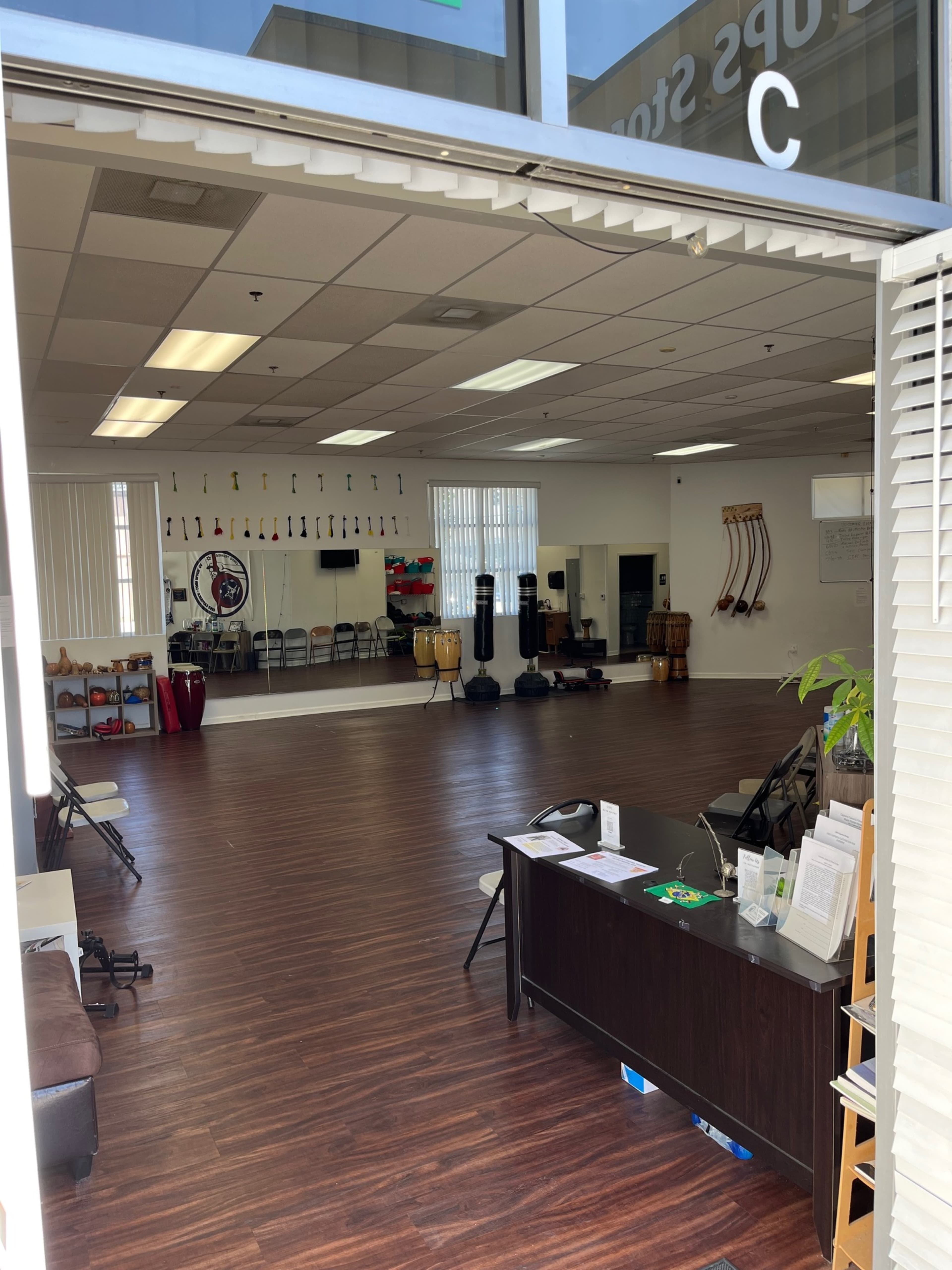 The image shows a spacious studio with wooden flooring, chairs arranged in rows, musical instruments on display, and a reception area with a desk and brochures.