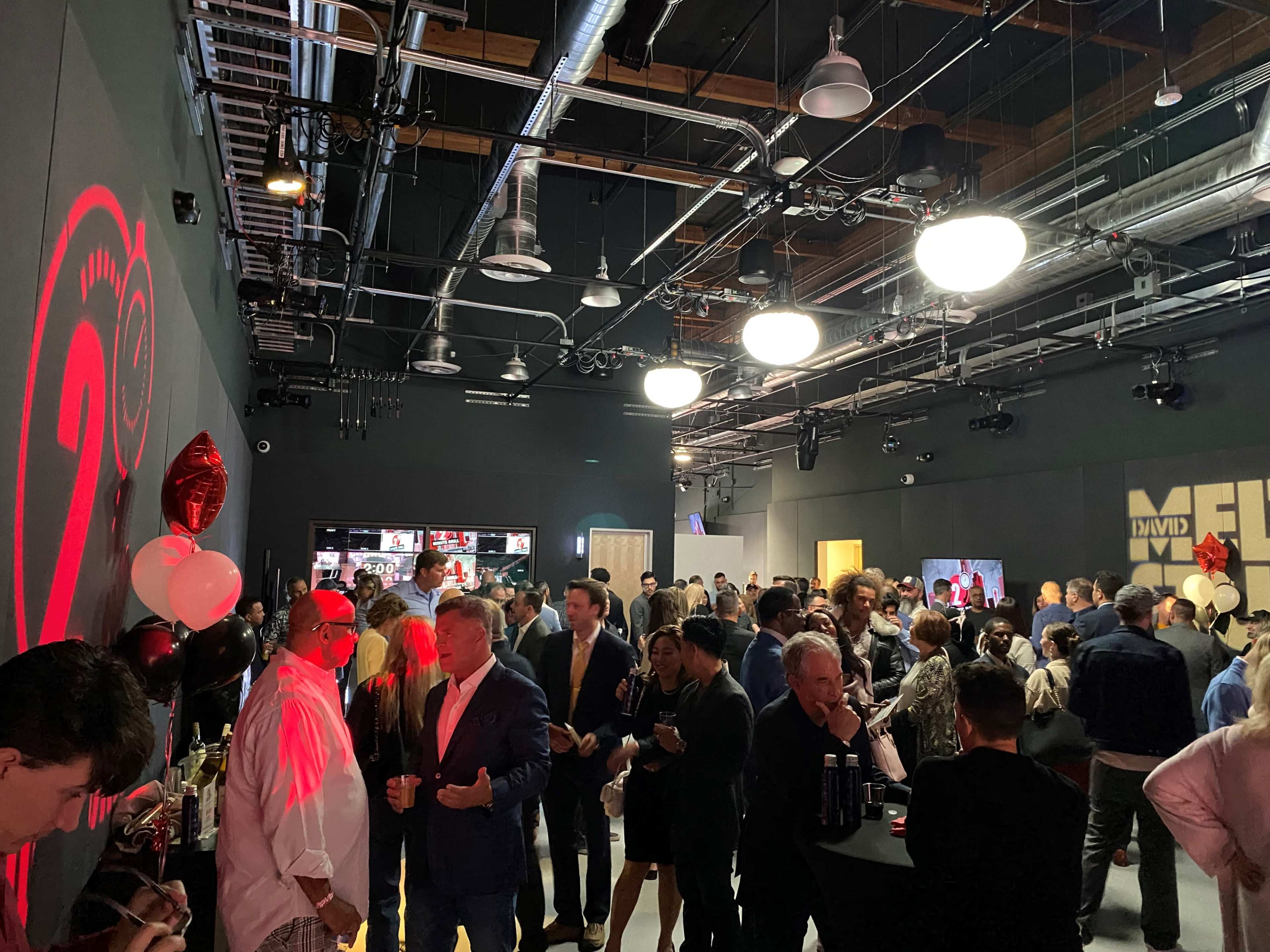 A crowded event space features guests mingling, with decorations including balloons and illuminated wall art.