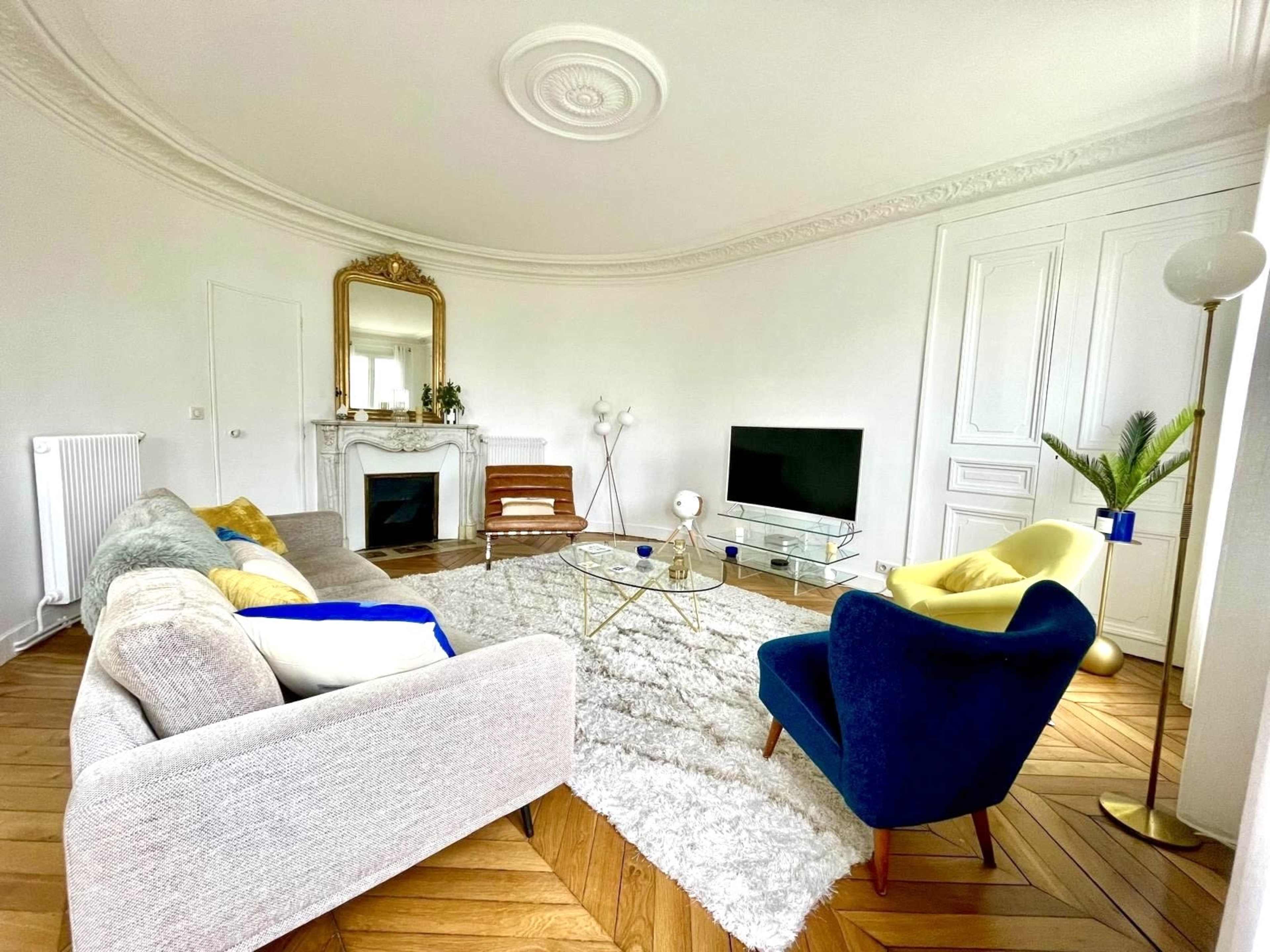 The living room features a light-colored sofa and a blue armchair, a coffee table, a large area rug, a flat-screen TV, and a decorative fireplace.