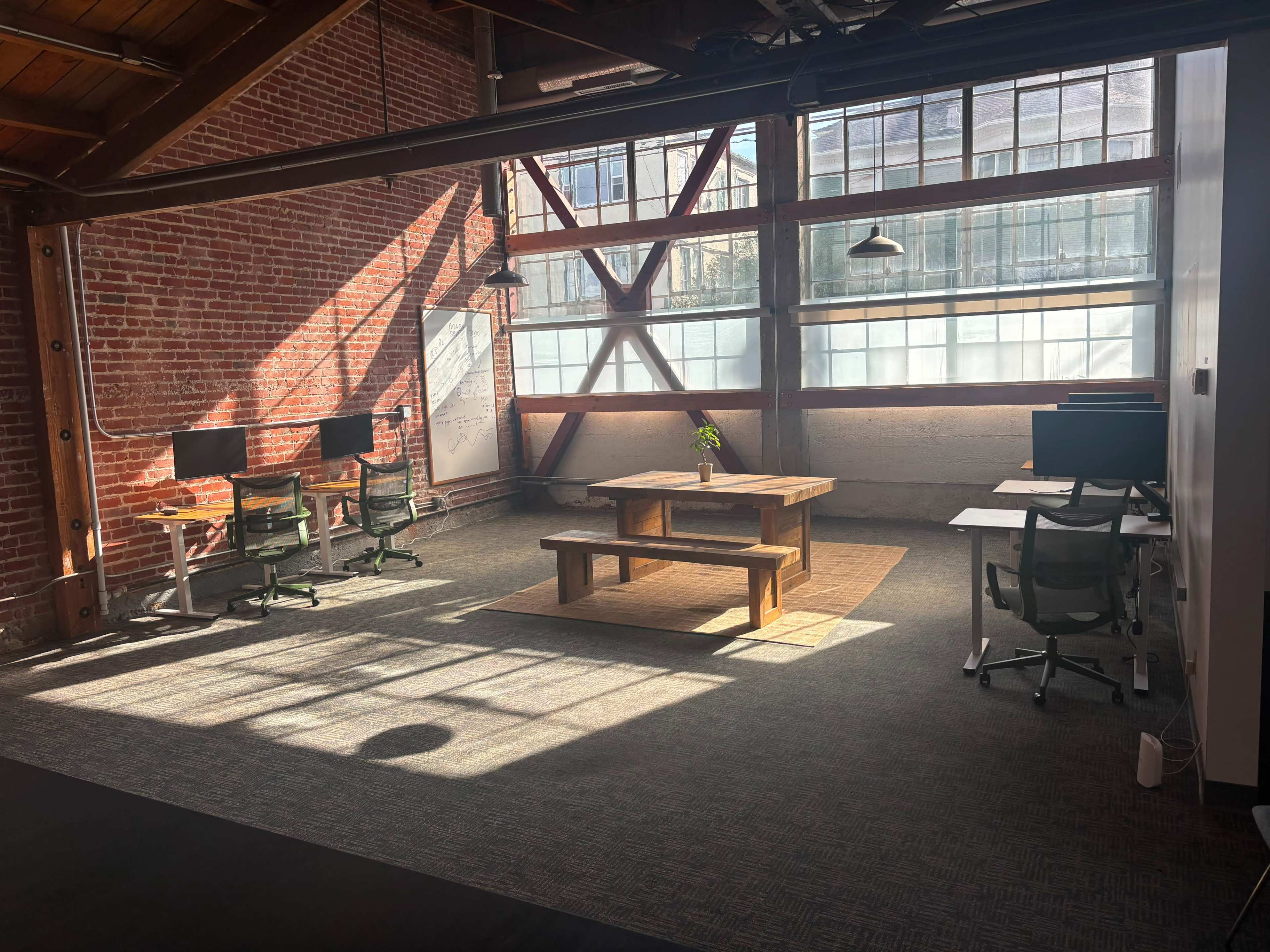 The image shows a spacious office interior with exposed brick walls, a large wooden table in the center, and desks with computers arranged along the sides, illuminated by sunlight streaming through large windows.