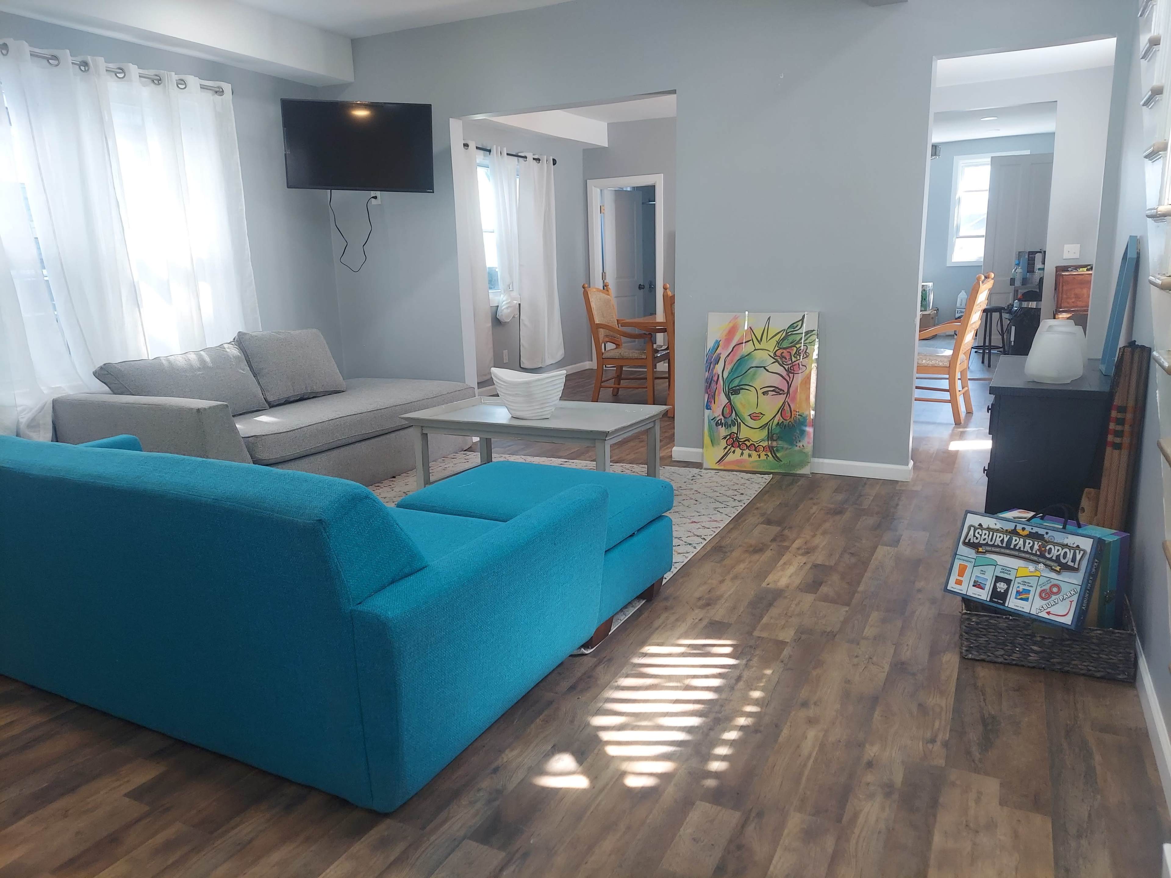 A bright living room with blue and gray furniture, hardwood floors, a television on the wall, and a colorful art piece on display.