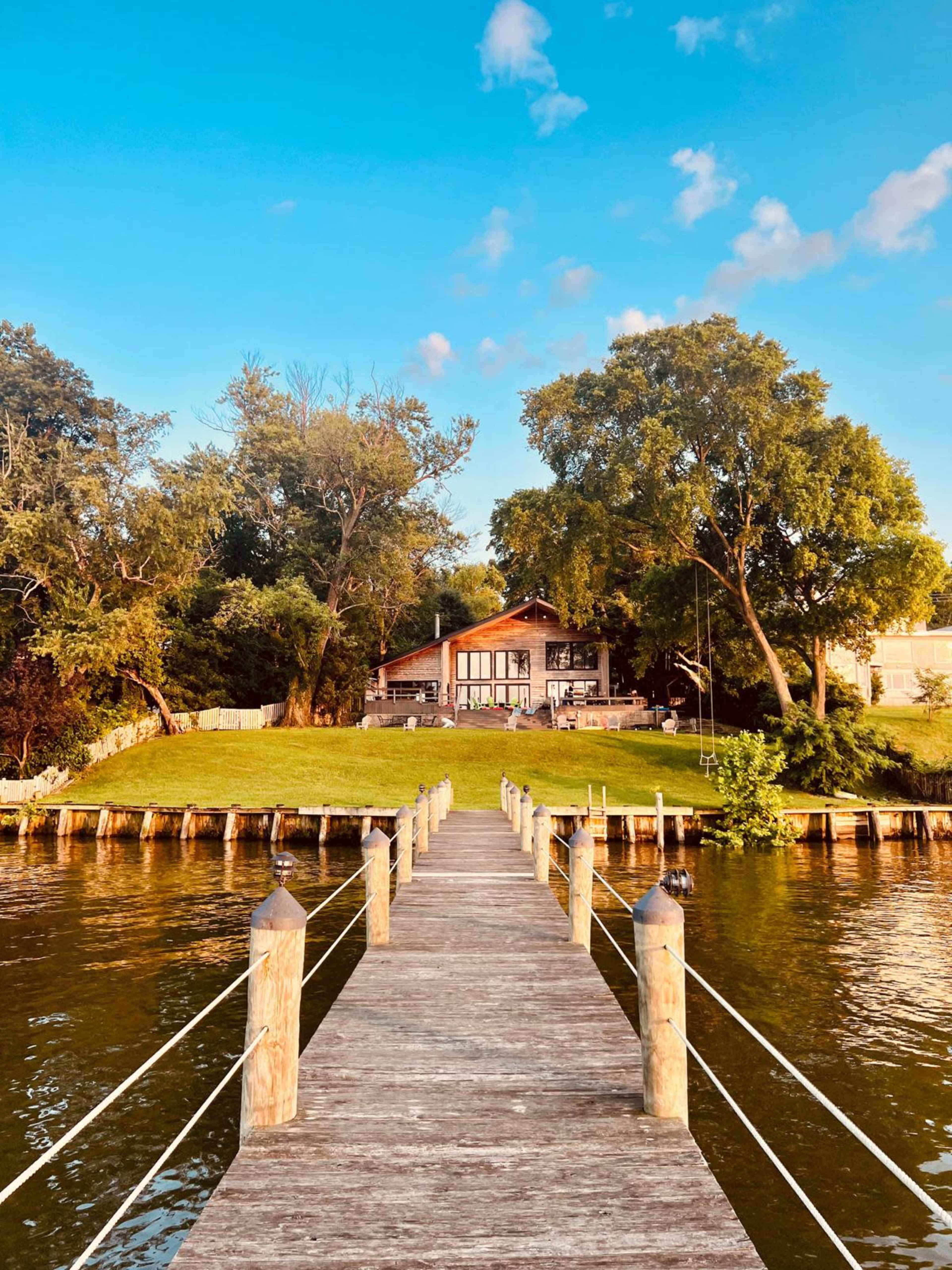 A wooden dock extends into a still body of water, leading to a house set back on a green lawn surrounded by trees.