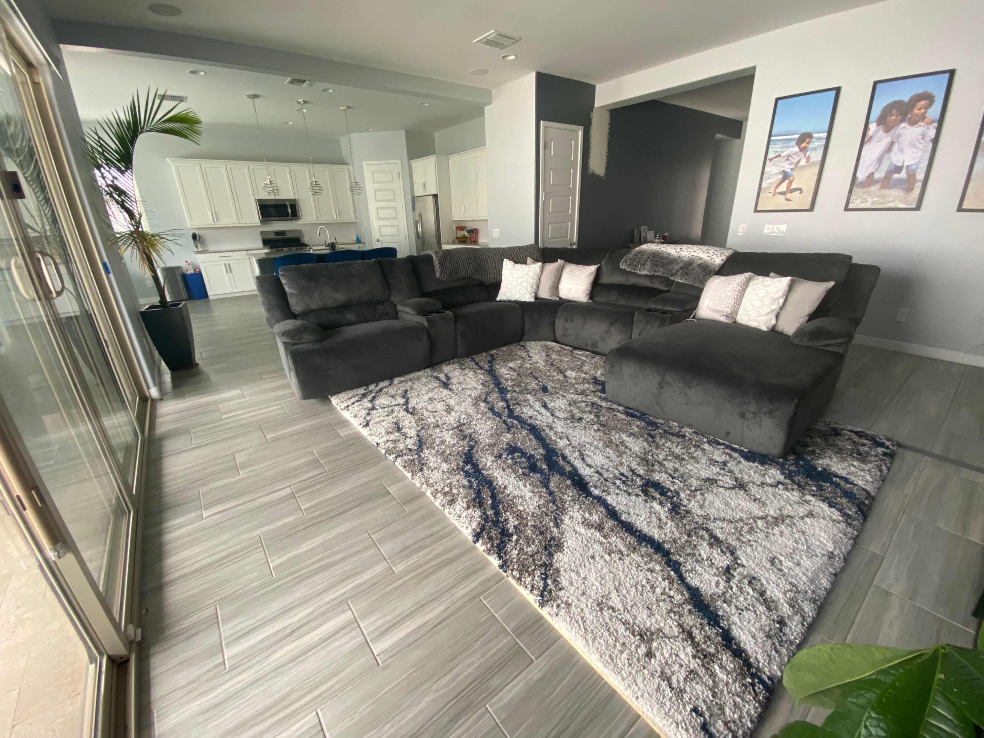 A modern living room features a gray sectional sofa, a patterned area rug, and framed photographs on the wall, with an open kitchen visible in the background.