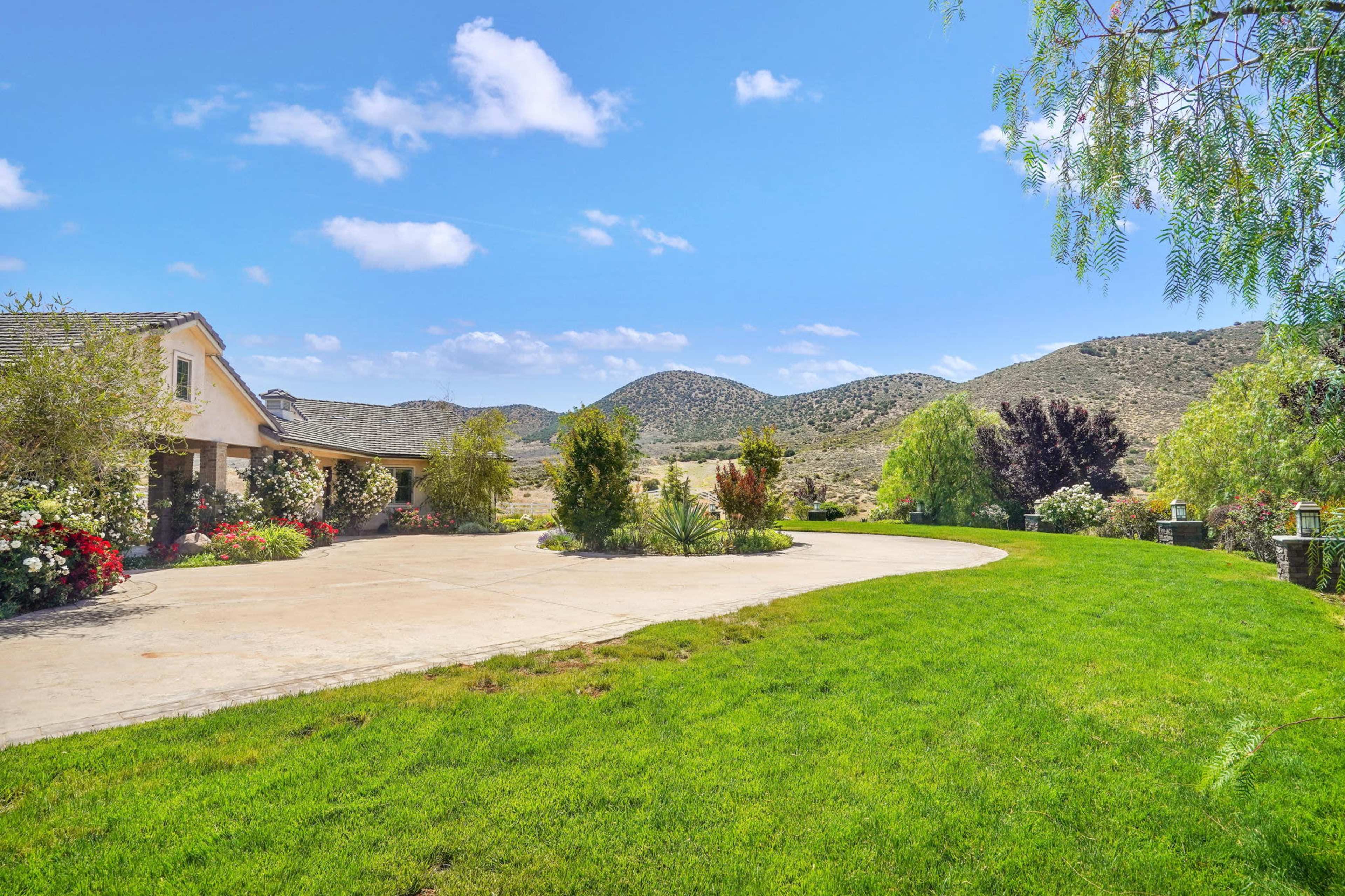 A landscaped yard with a curved driveway, surrounded by mountains and greenery under a clear sky.