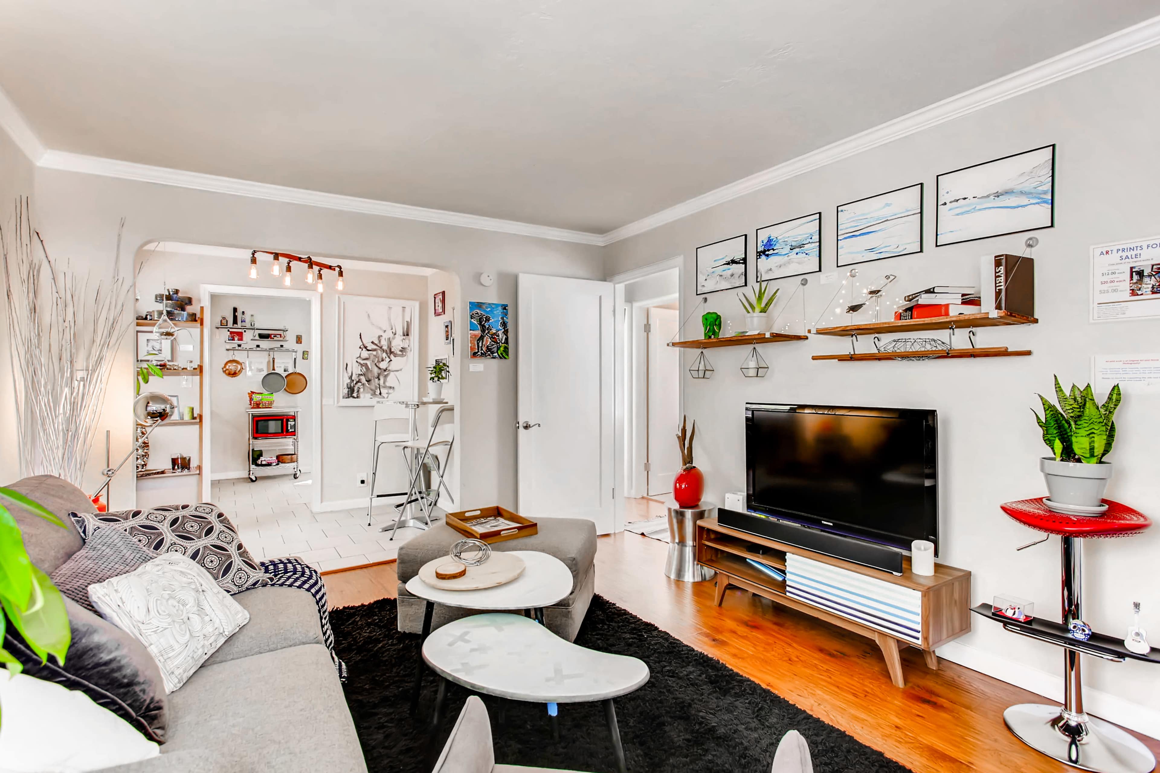 The image shows a modern living room with a gray sofa, a wall-mounted television on a wooden console, and decorative shelves displaying plants and artwork.