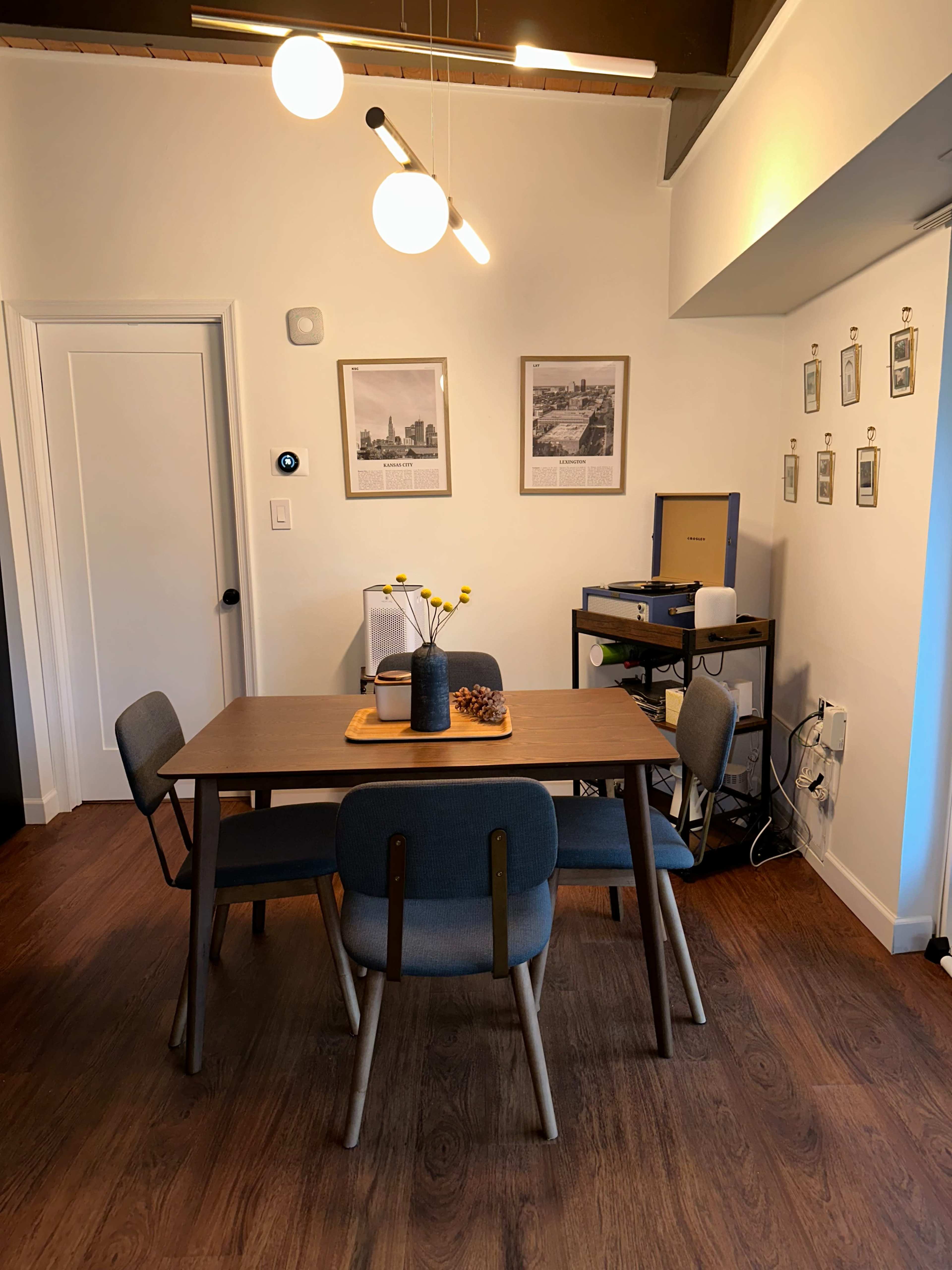 A dining area features a wooden table surrounded by four upholstered chairs, with framed pictures on the walls and a small shelf displaying a record player.