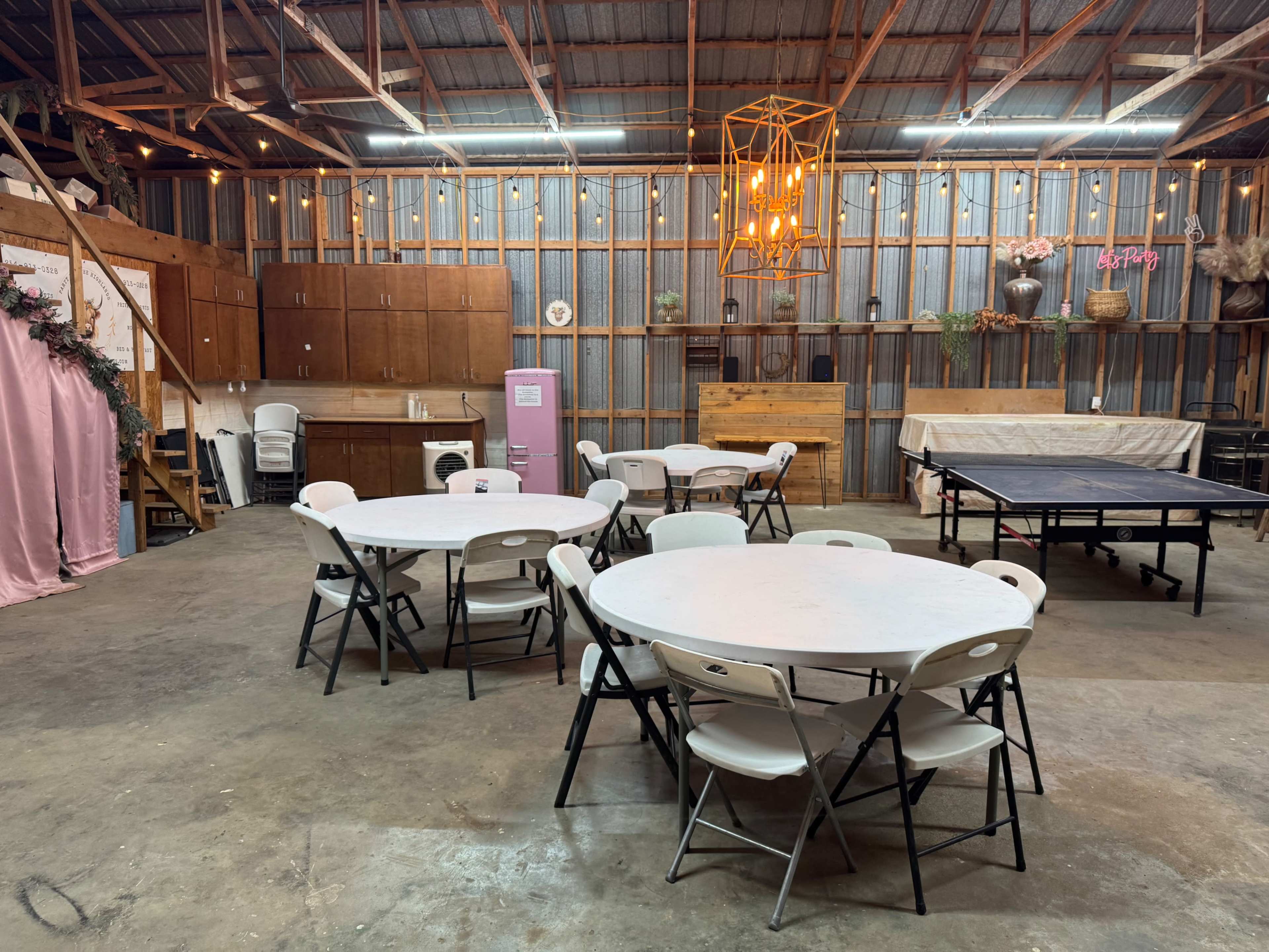 The image shows an interior space with several round tables surrounded by chairs, a ping pong table, wooden walls, and a decorative light fixture above.