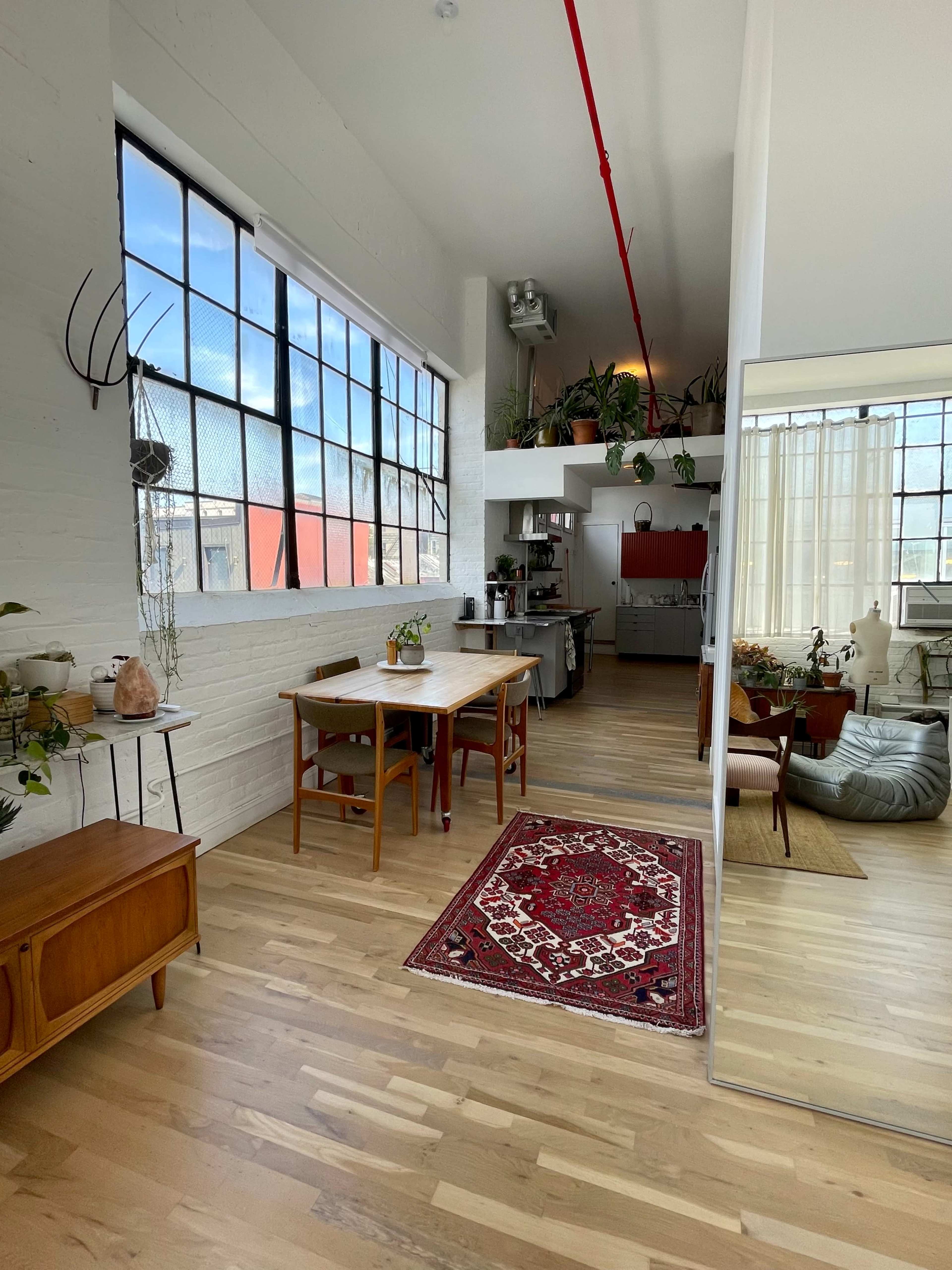 The image shows an open-concept living space with a wooden dining table, a kitchenette in the background, and large windows allowing natural light to enter.