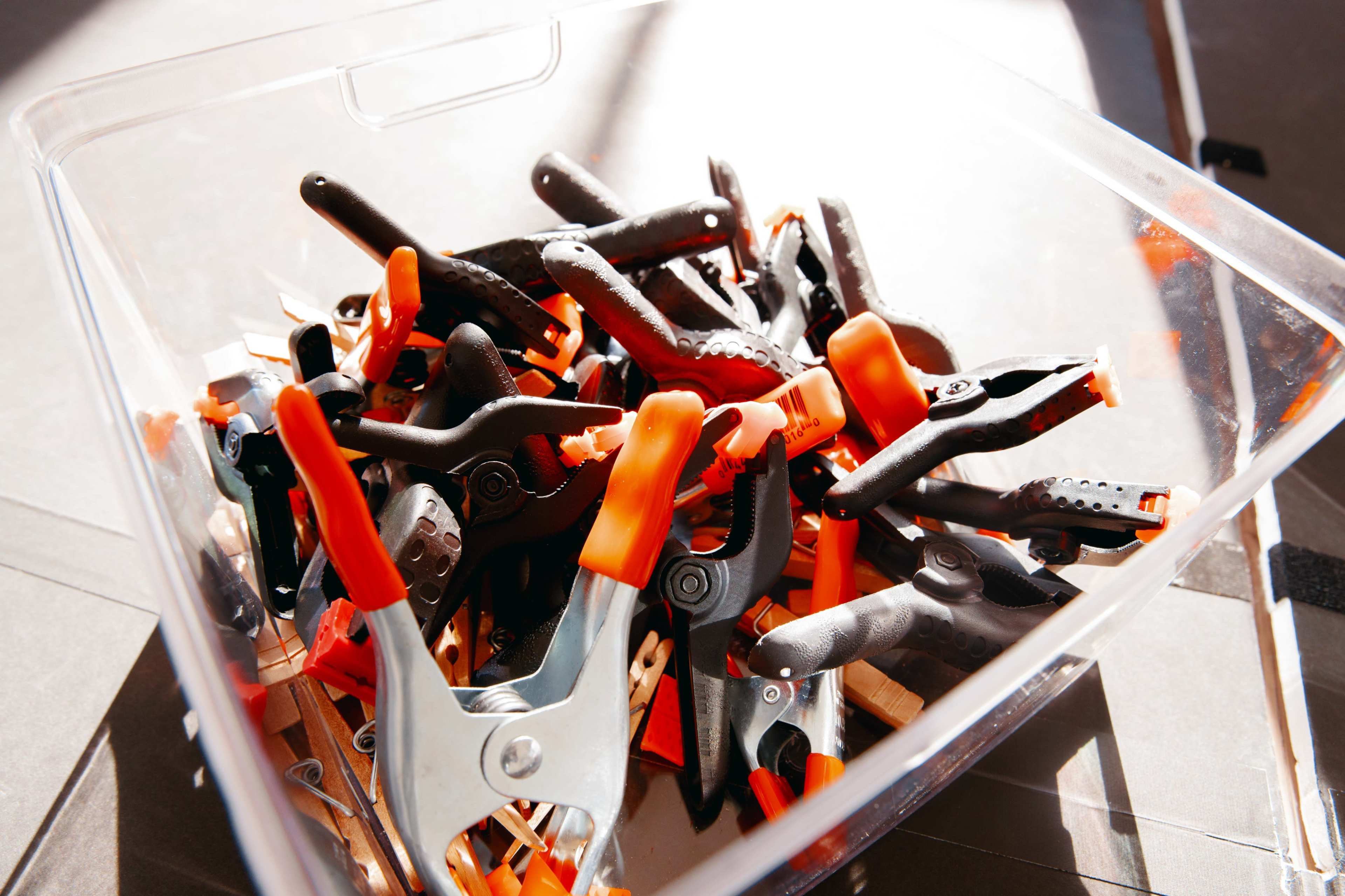 A clear container holds a variety of black and orange spring clamps.