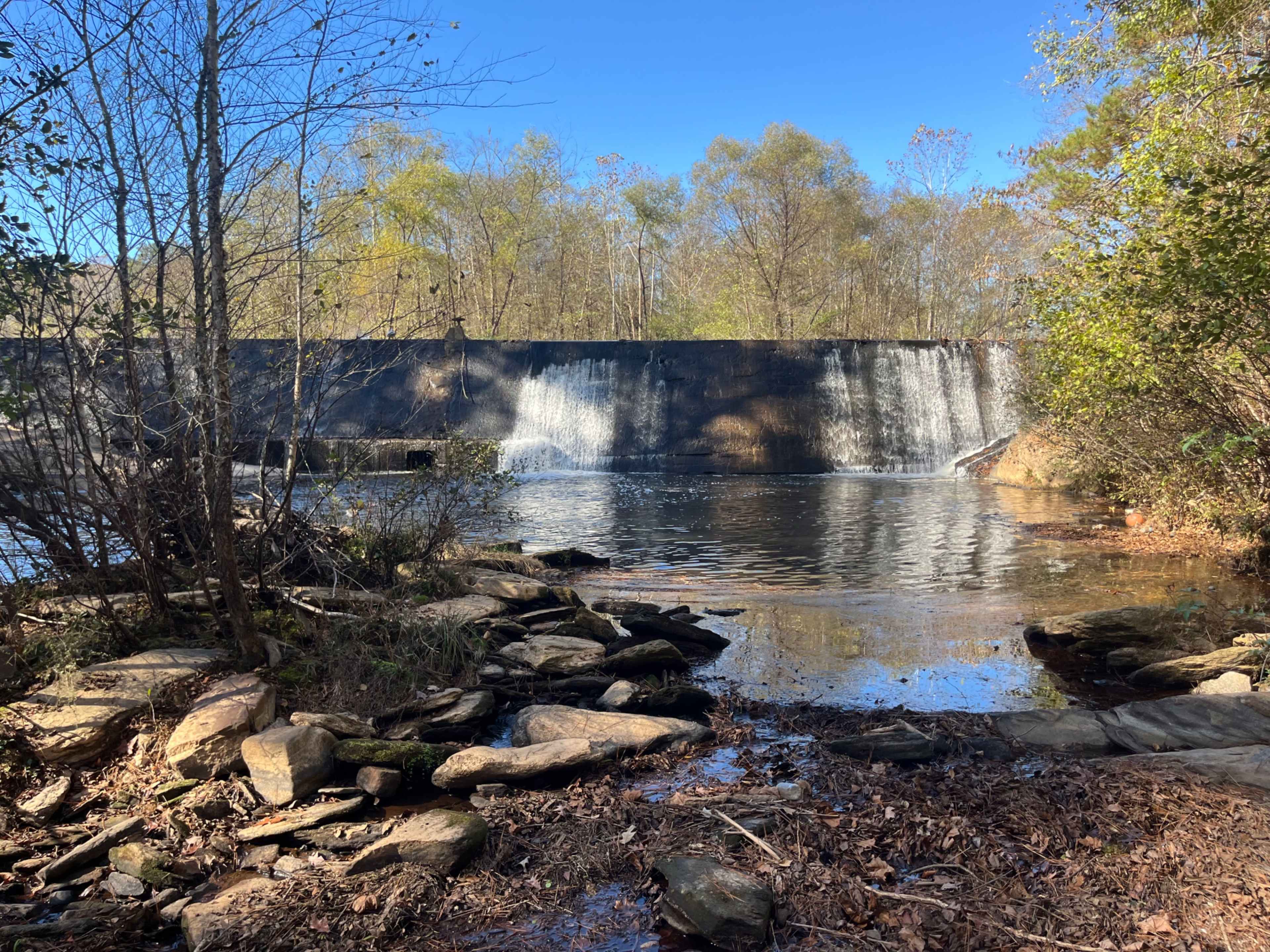 A tranquil scene shows a waterfall cascading over a dam into a calm body of water, surrounded by trees and rocky shorelines.