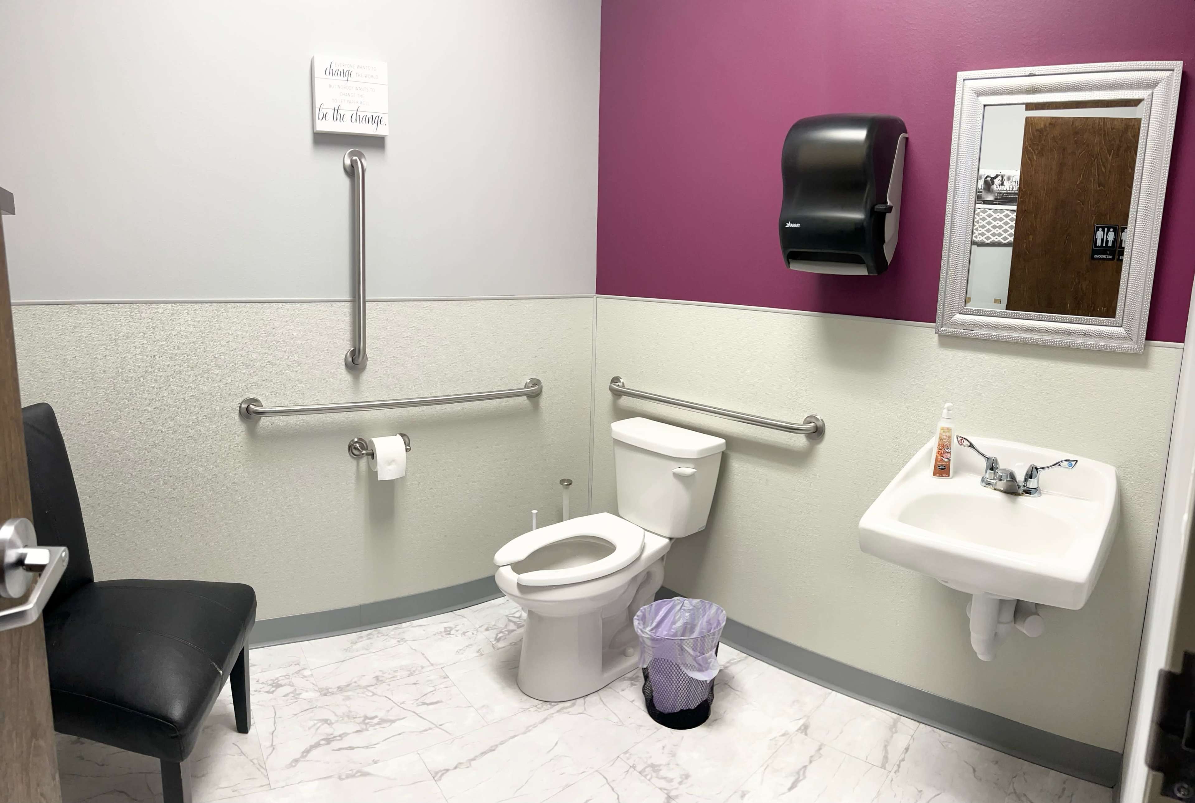A bathroom equipped for accessibility, featuring a toilet, a sink, grab bars, and a chair against the wall.