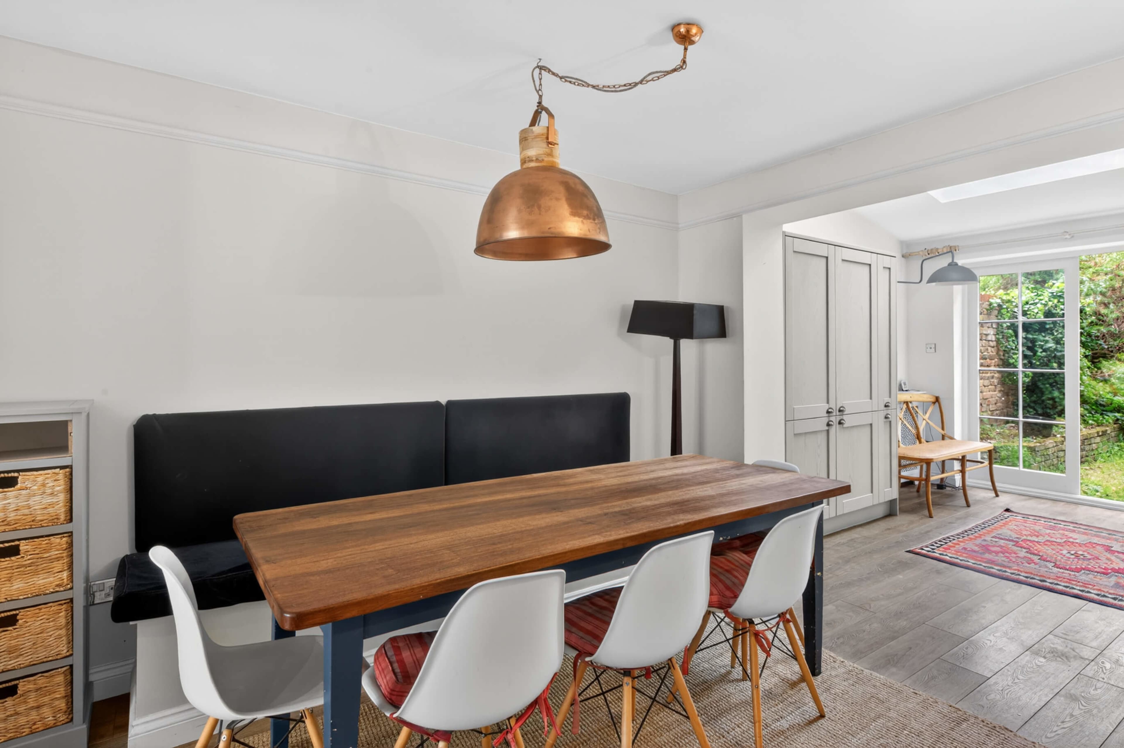 The image shows a dining area with a wooden table surrounded by white chairs, a copper pendant light overhead, and a door leading to an outdoor space.