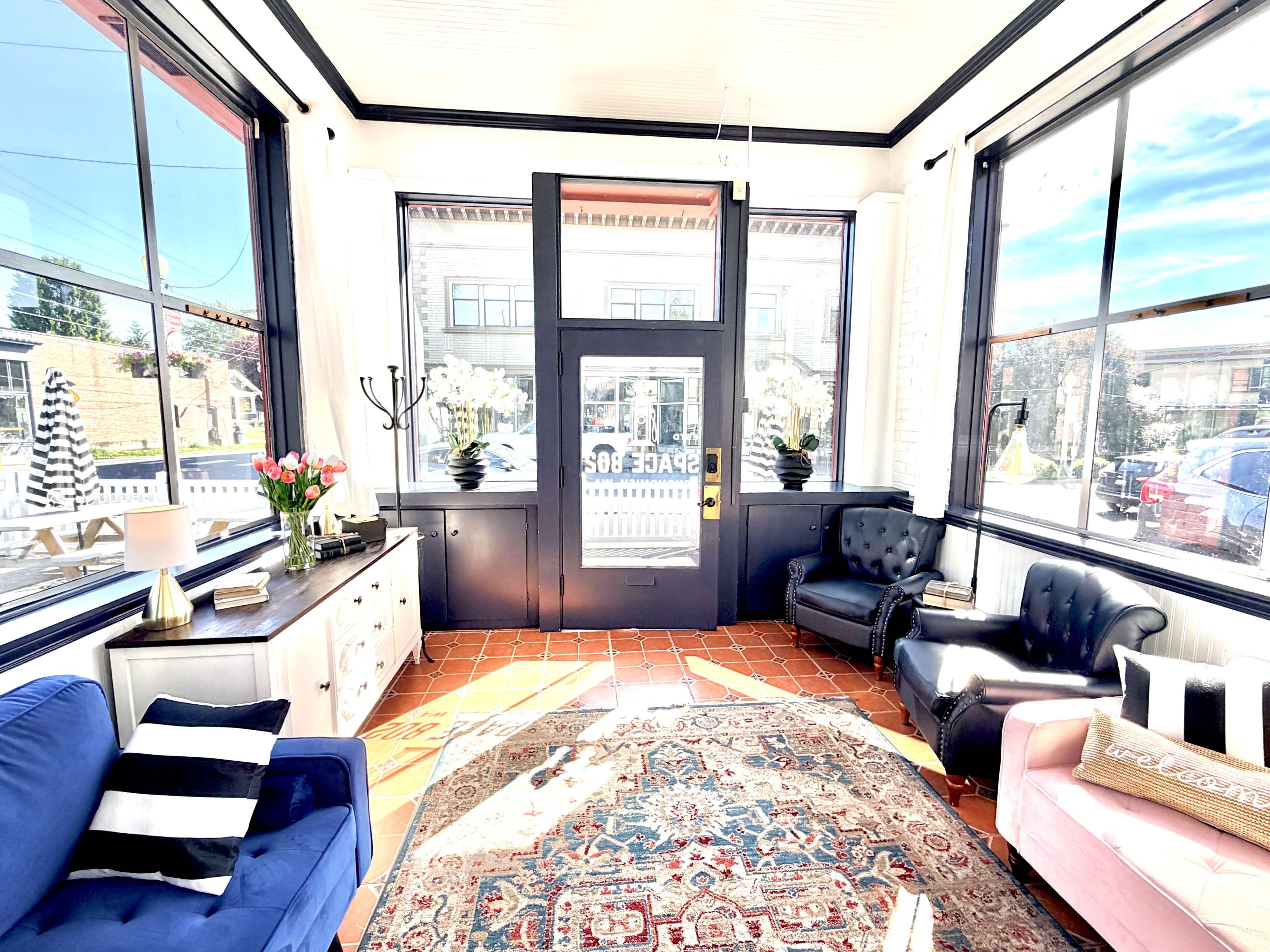 The image shows a bright sunlit area with large windows, featuring a mix of seating including a blue couch, a pink armchair, and a patterned rug on a tiled floor.