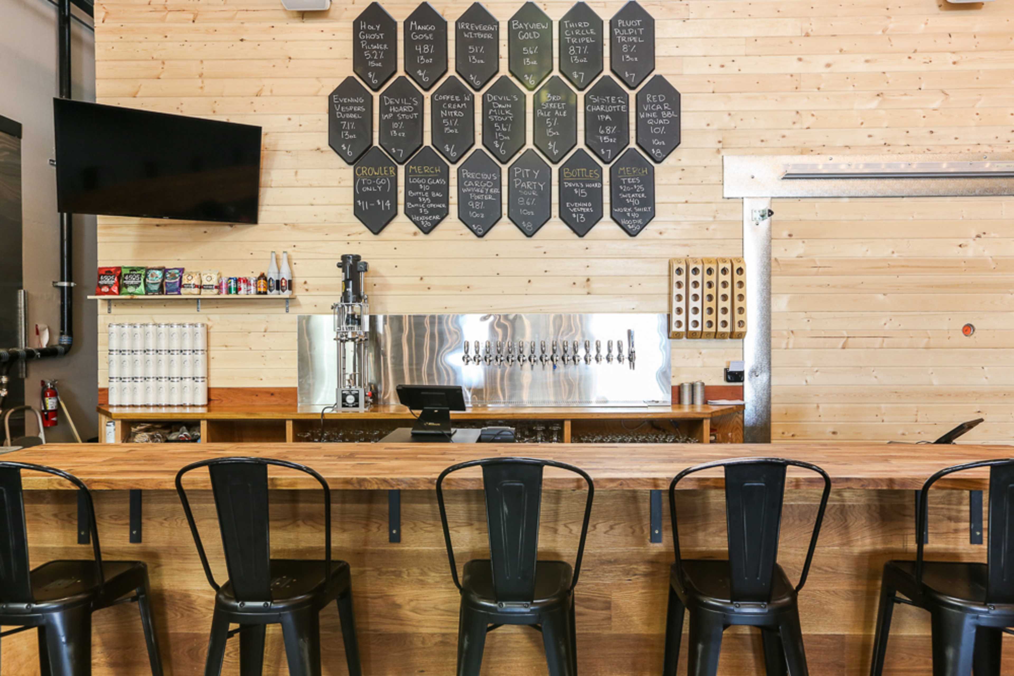 A wooden bar with metal stools faces a wall featuring a drink menu and multiple beer taps.