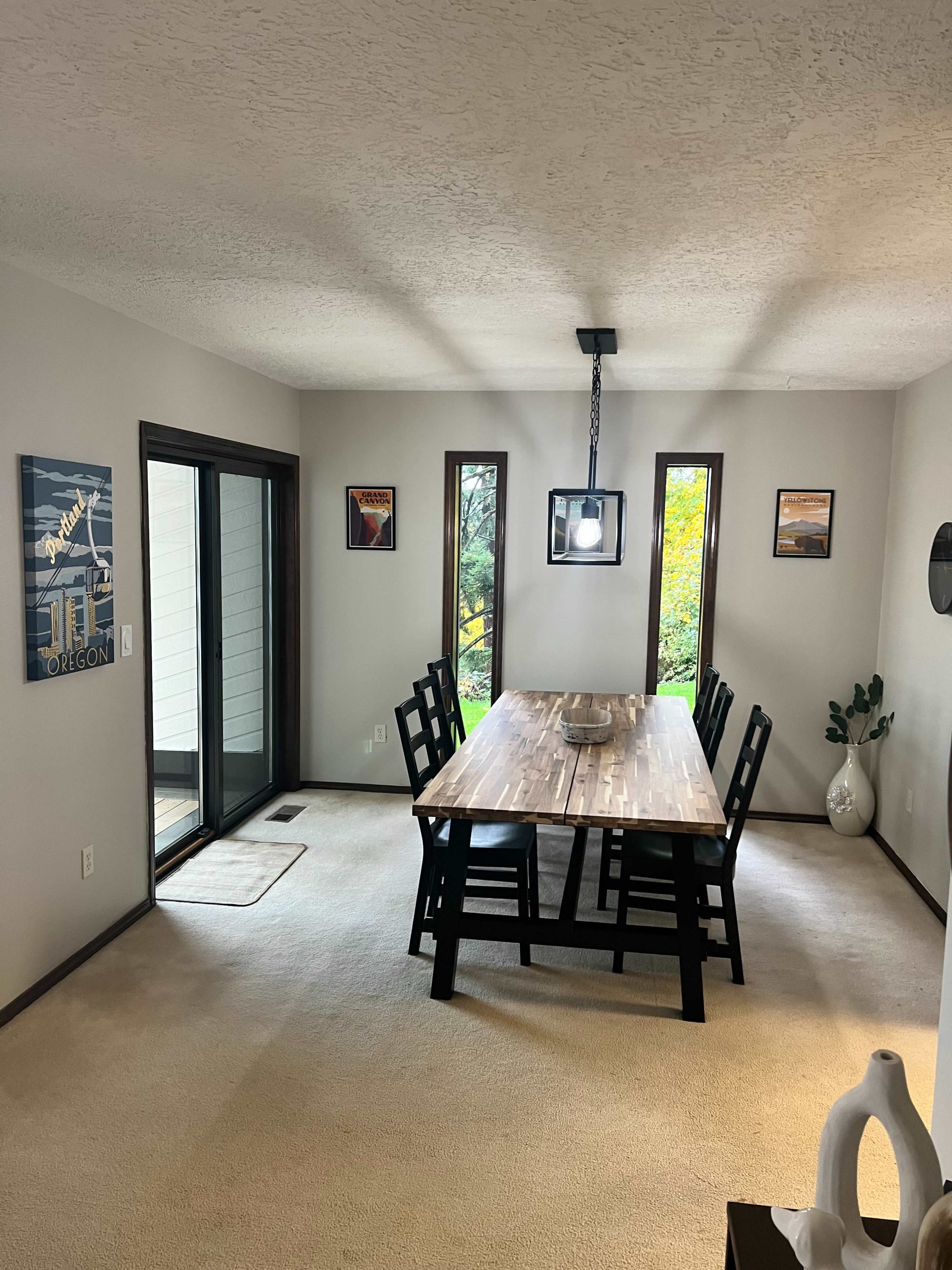 The image shows a dining room with a wooden table and six black chairs, flanked by three vertical windows and a sliding glass door, creating an open and spacious feel.