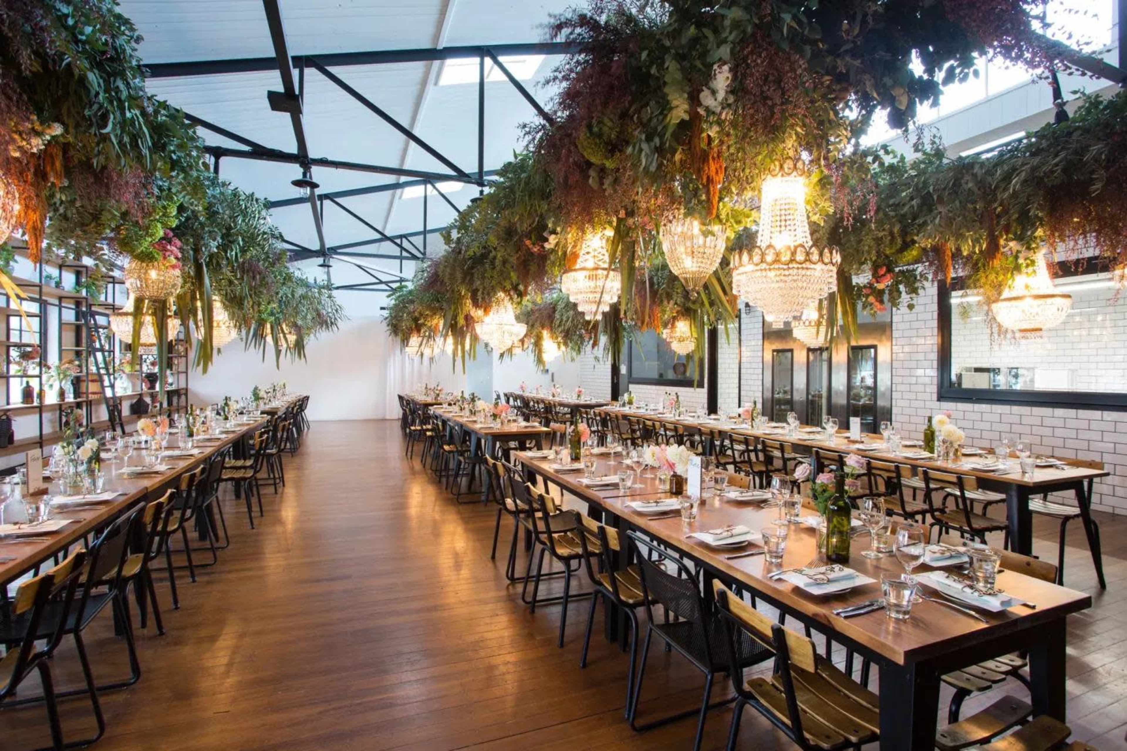 A spacious dining area with long tables set for a meal, surrounded by hanging greenery and decorative chandeliers.