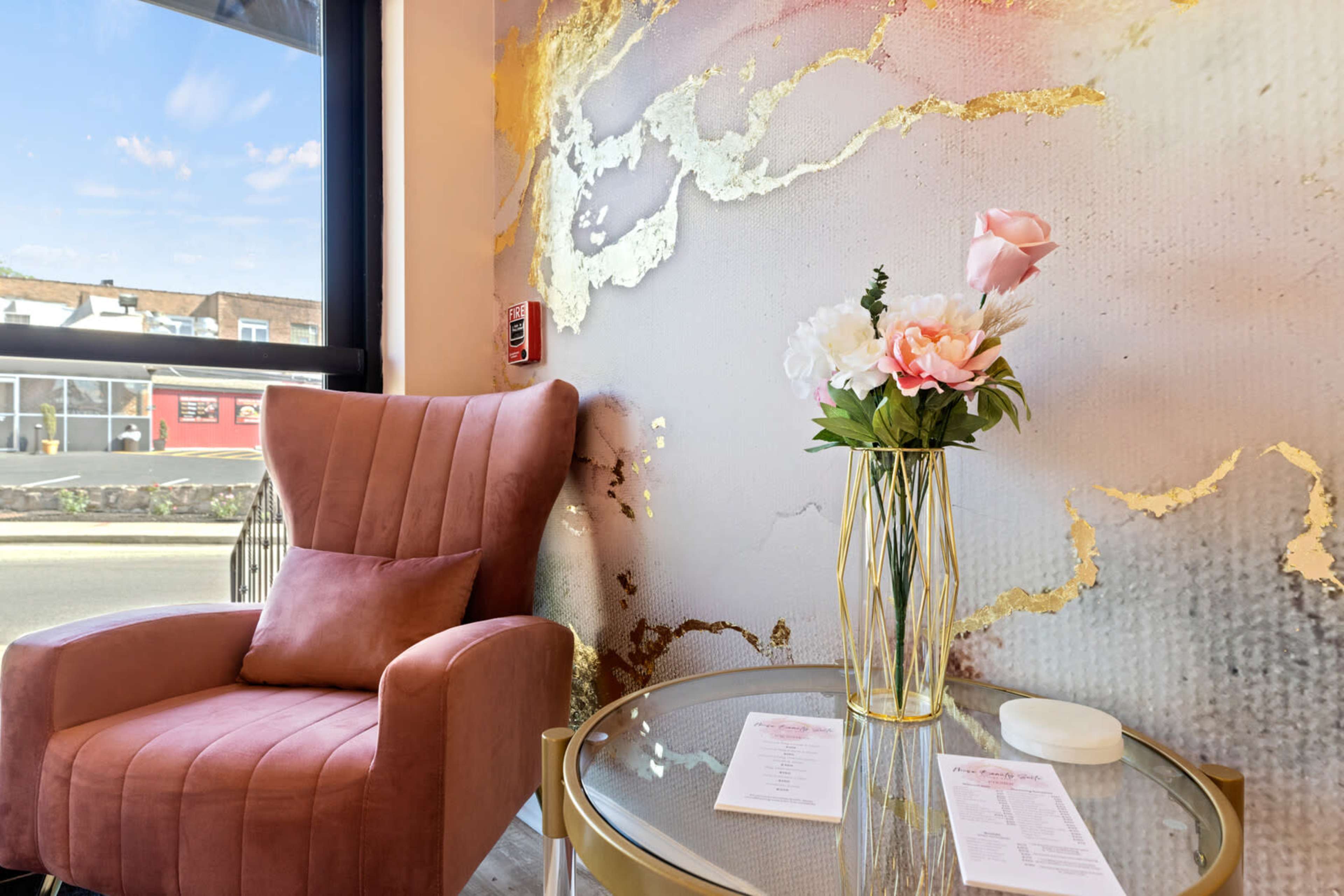 A pink velvet armchair sits beside a gold-accented table topped with a floral arrangement and menus, against a backdrop of a decorative wall featuring a marbled design with gold accents.