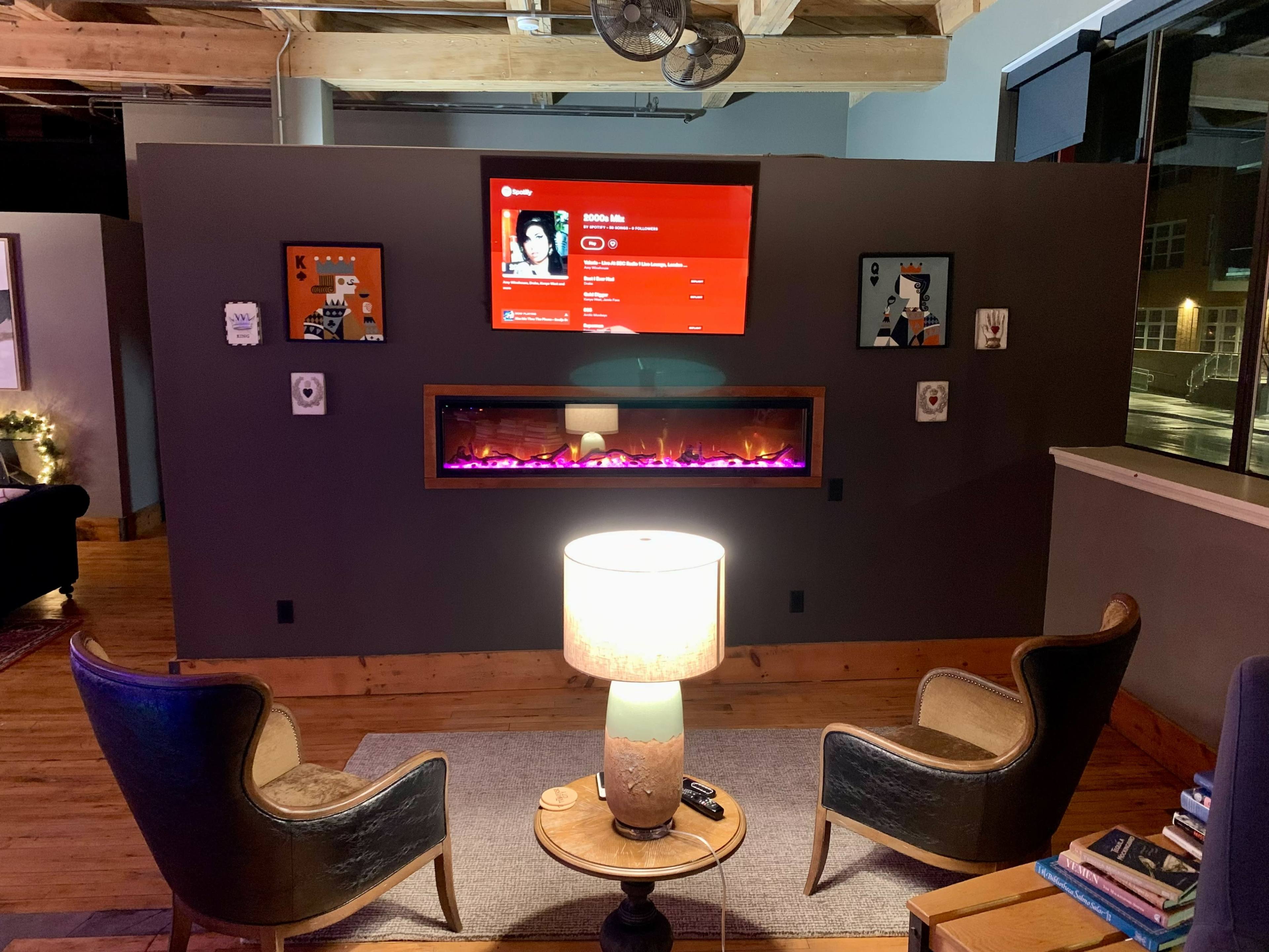 A living room area with two chairs facing a wall-mounted TV above an electric fireplace, accompanied by a lamp and various decorative items.