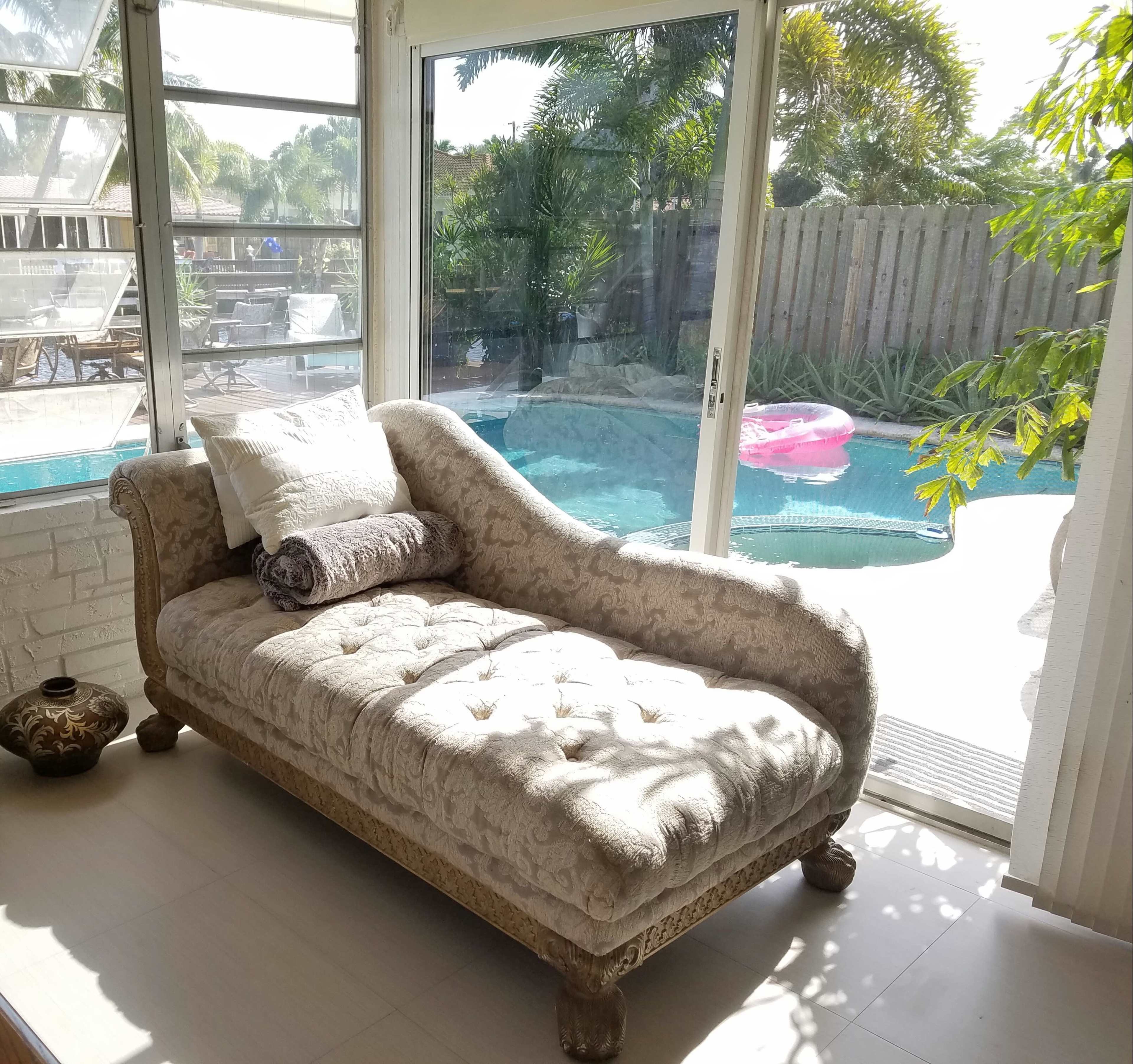 A chaise lounge with decorative cushions is positioned near a large window overlooking a swimming pool surrounded by greenery.