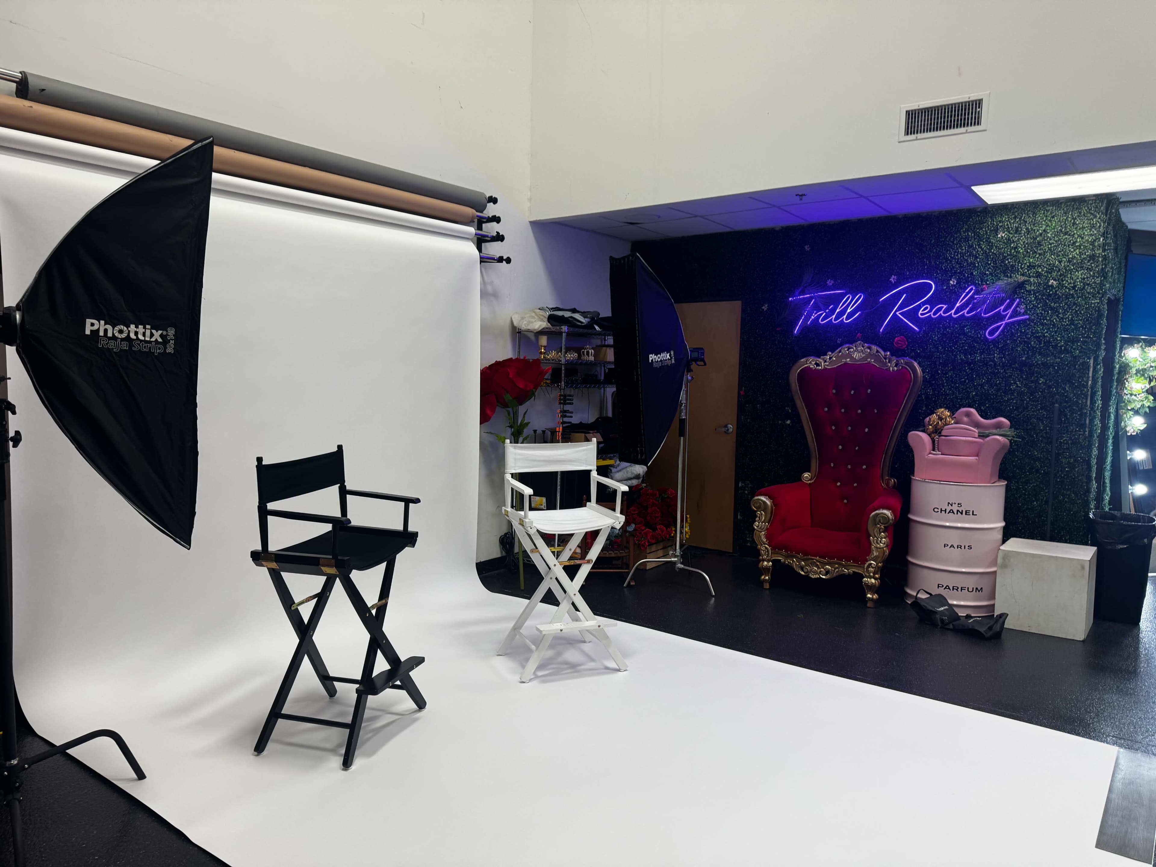 The image shows a photo studio setup featuring two director's chairs, a large ornate red throne, and a backdrop with white paper and decorative greenery.