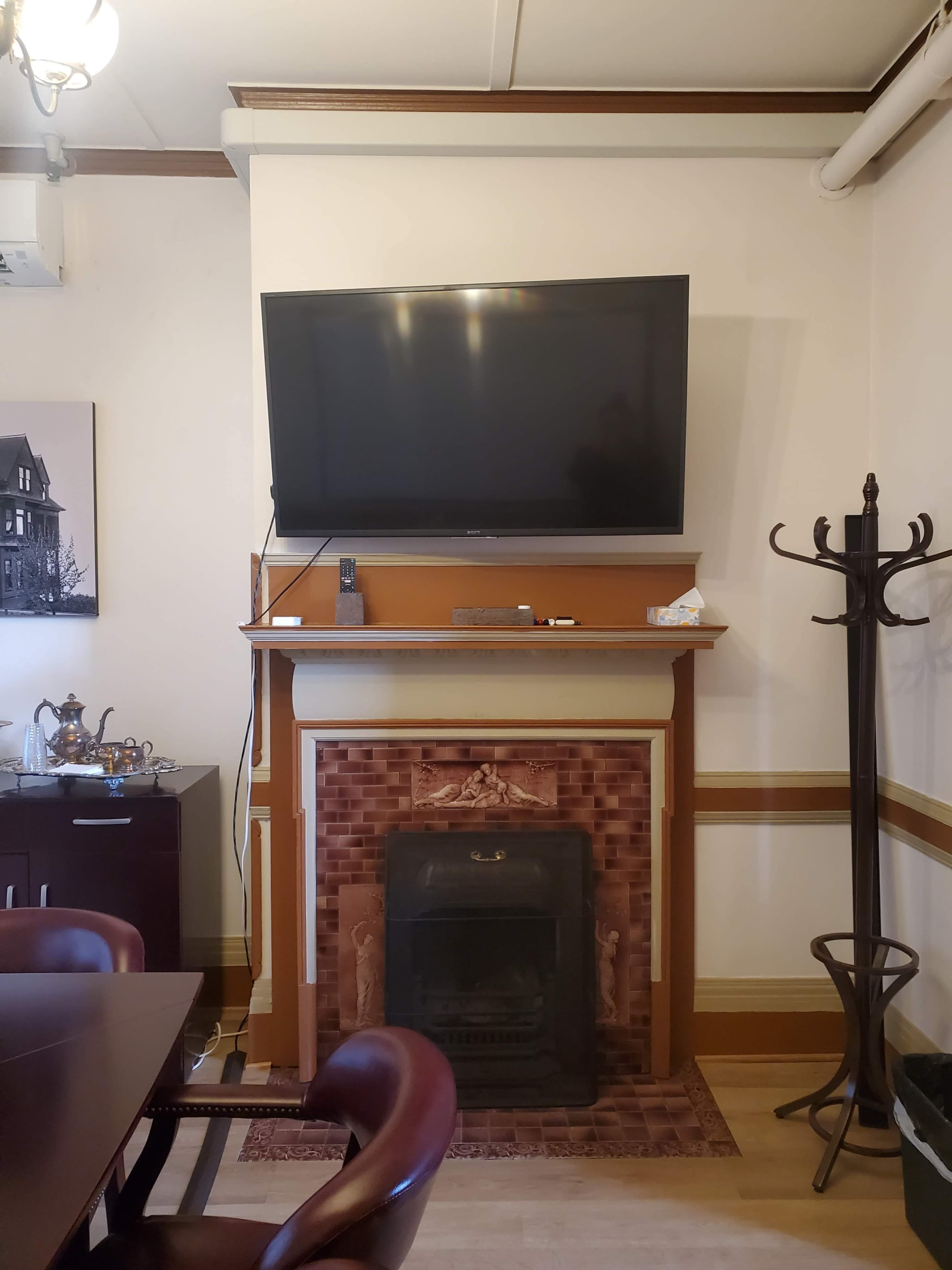 A living room with a mounted television above a decorative fireplace, accompanied by a wooden dining table and a coat rack.