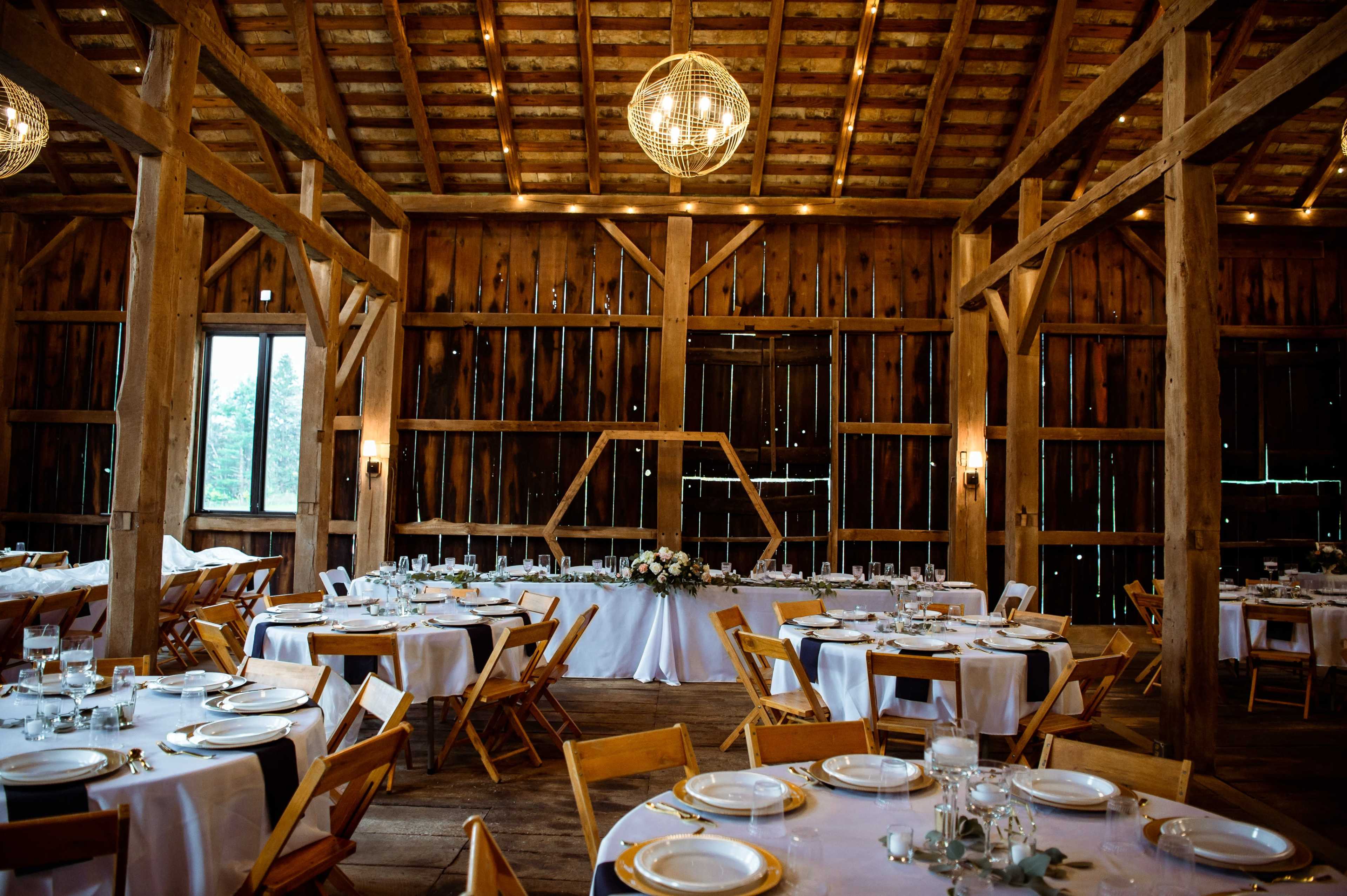A rustic barn interior set up for a wedding, with round tables covered in white tablecloths and wooden chairs arranged around them.