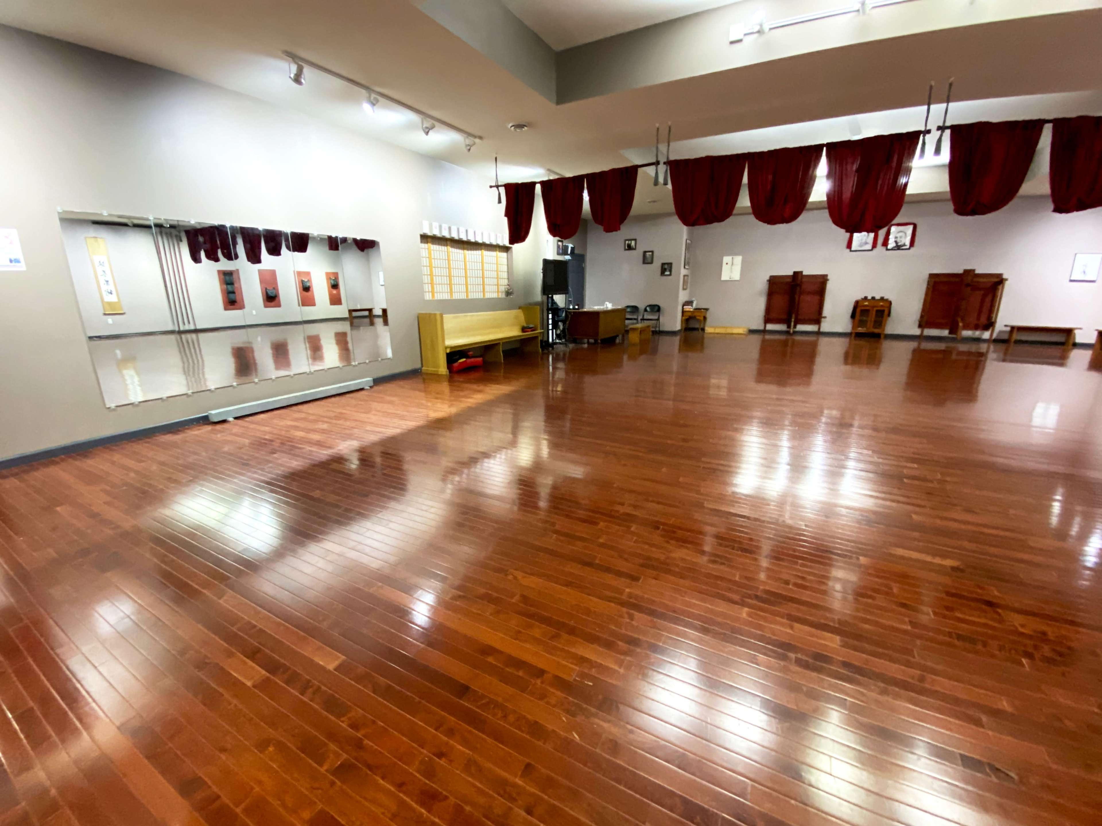 The image shows a spacious wooden-floored studio with mirrors on one wall and red curtains hanging from the ceiling.