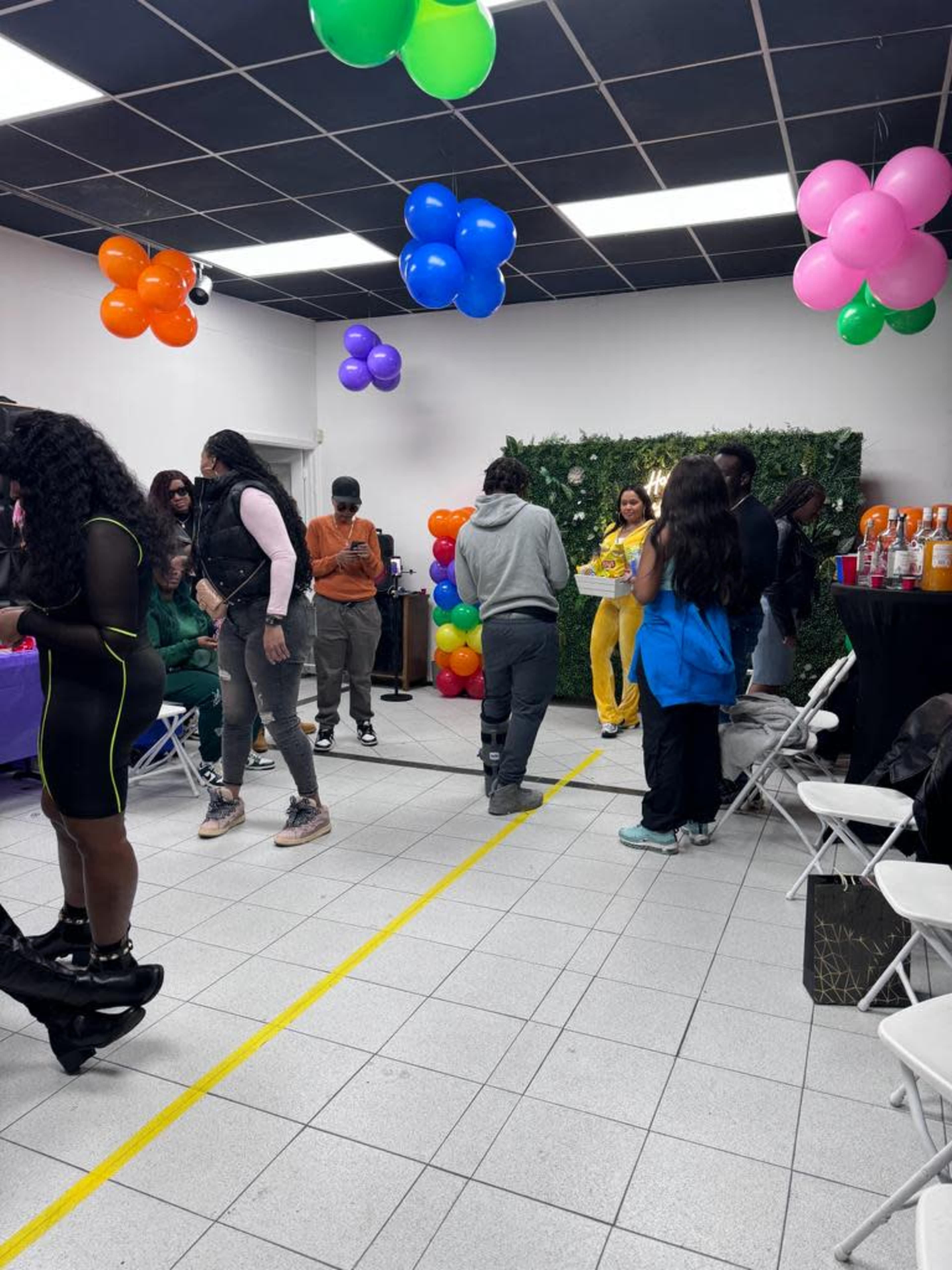 The scene shows a lively indoor party with guests mingling around colorful decorations and a table filled with drinks.