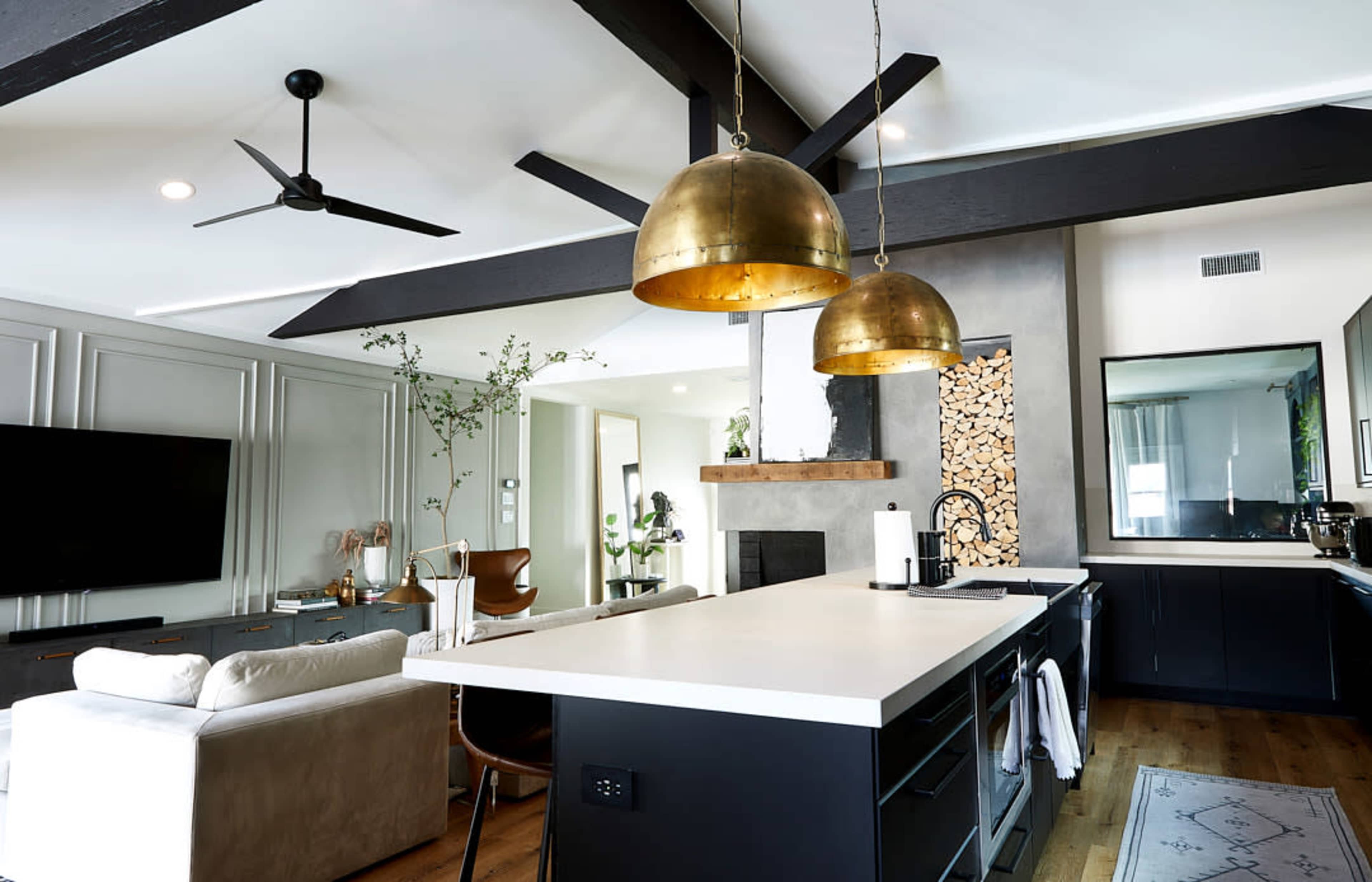 A modern kitchen and living room area featuring a large island with a white countertop, two brass pendant lights, and a wooden beam ceiling.