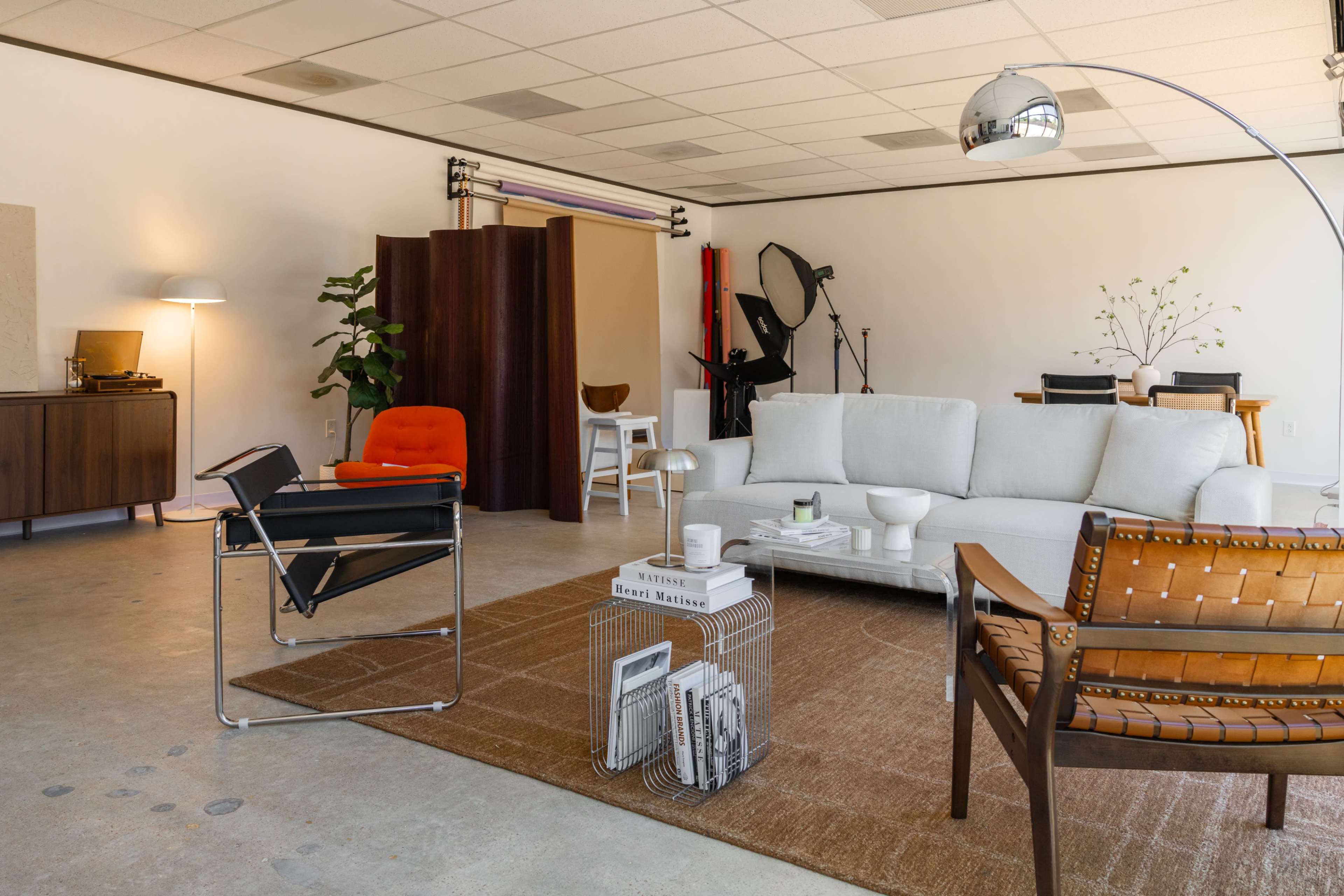 The image shows a modern, spacious interior with a light-colored sofa, an orange chair, a wooden dining table, and various photography equipment in the background.