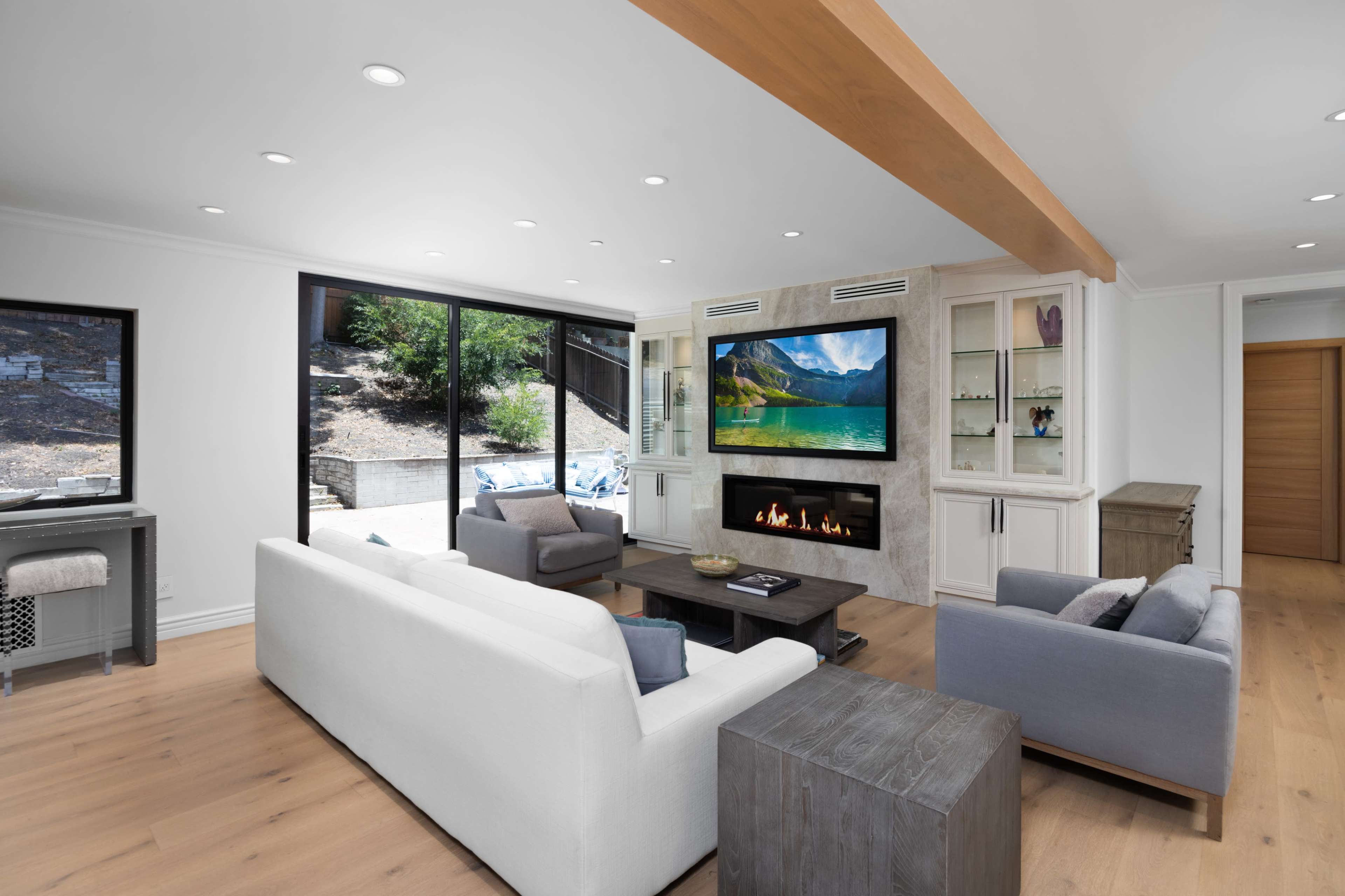 The living room features white and gray sofas arranged around a coffee table, with a modern fireplace and a large television set into the wall, and large windows providing a view of the outdoors.